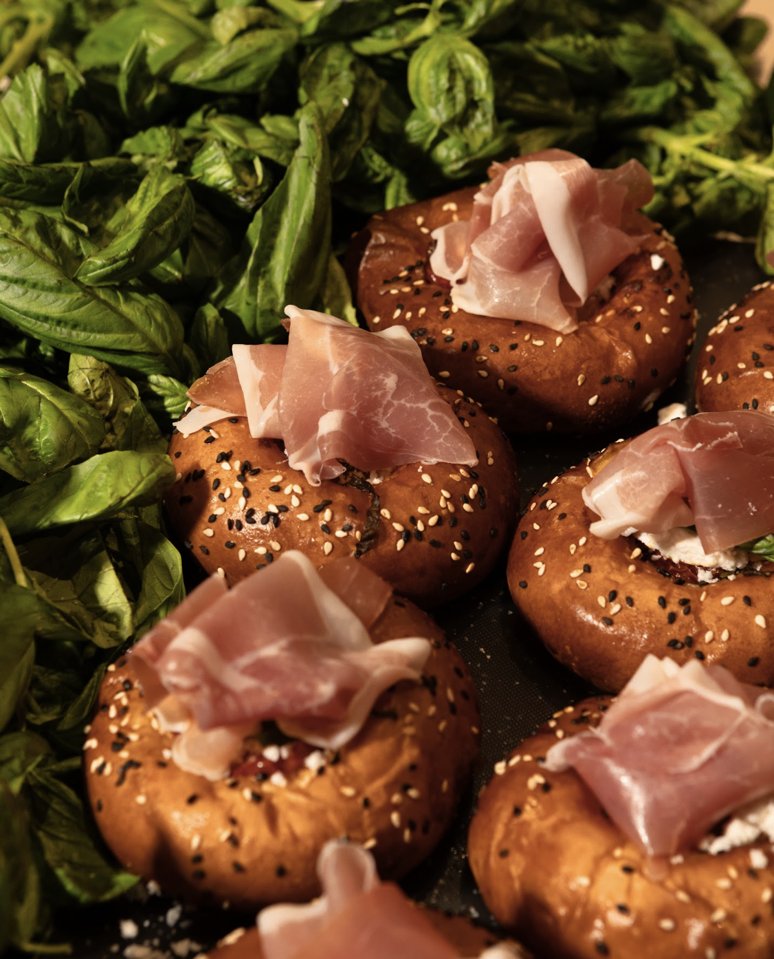 Tray of baked bagel bites topped with thin slices of prosciutto, surrounded by fresh basil leaves.