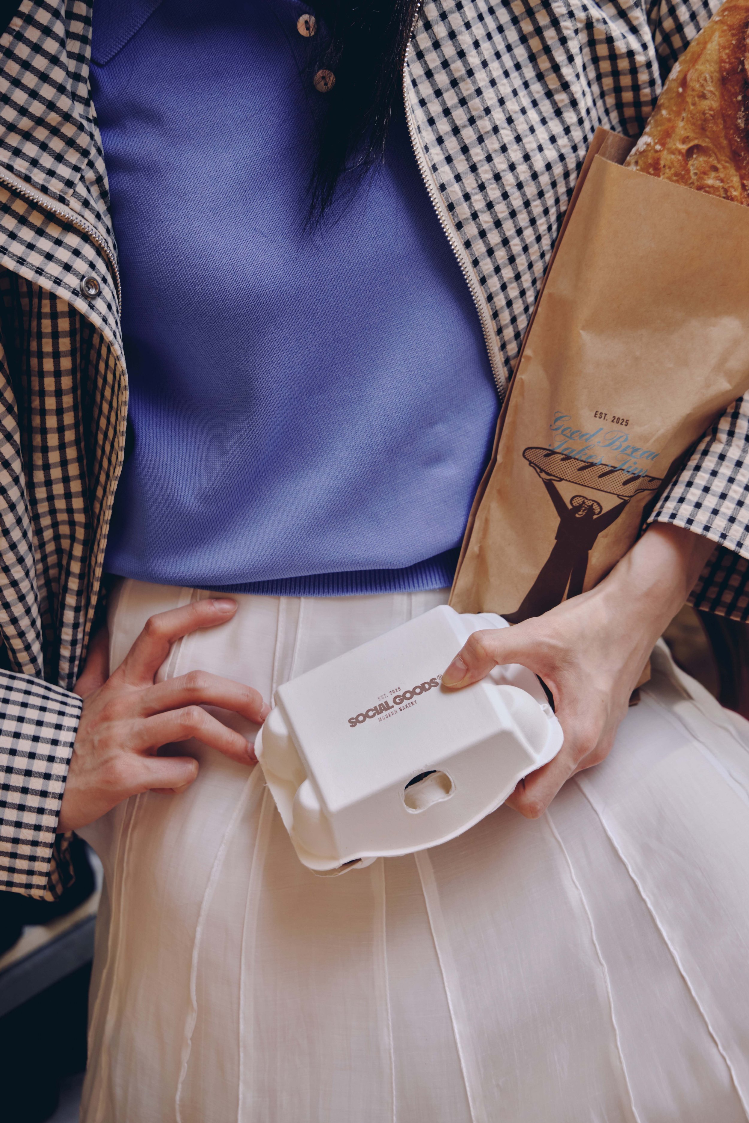 A customer in a checkered jacket carries a takeaway food container and bakery bag from Social Goods.