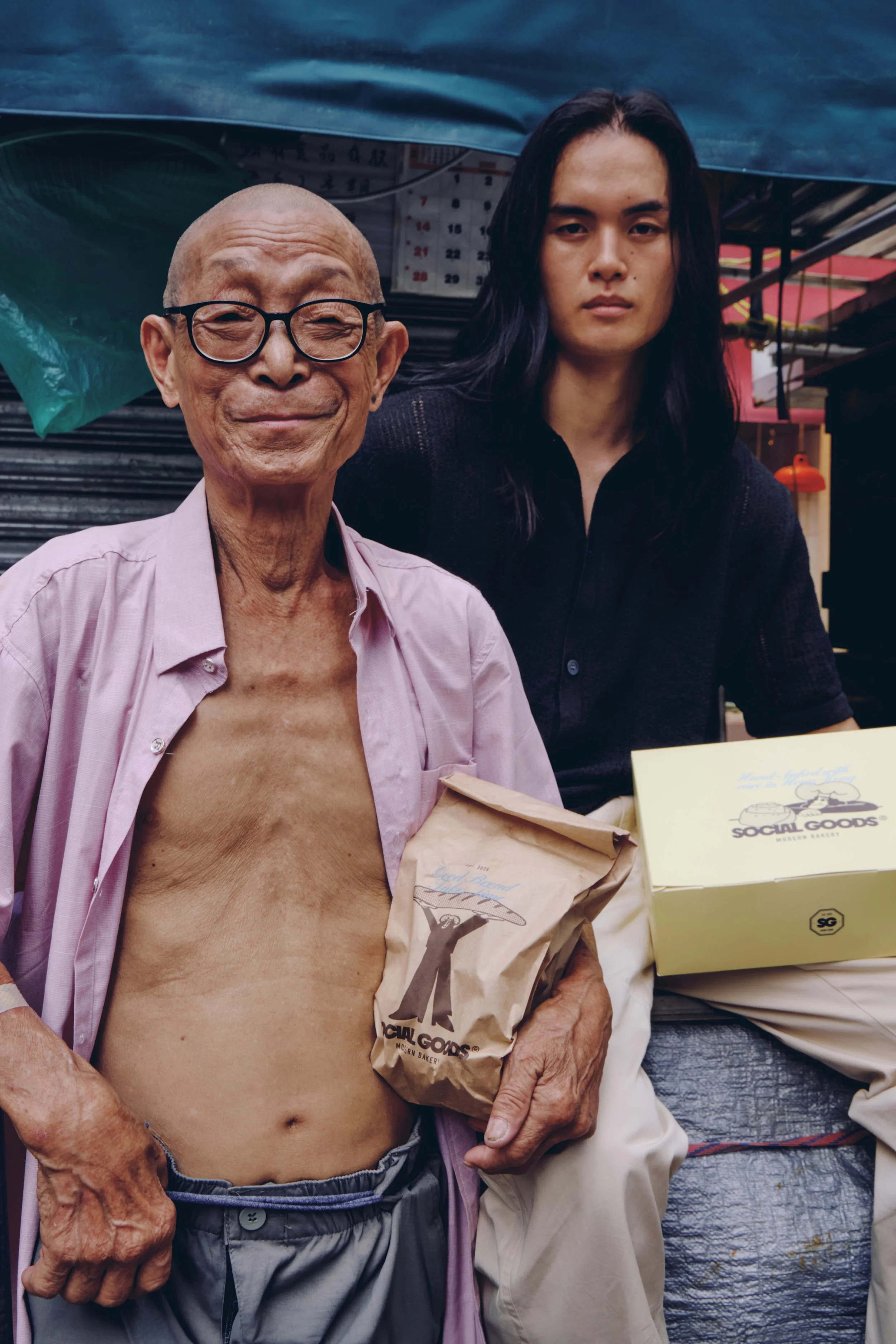 An elderly man and a young woman stand together outdoors near market stalls and umbrellas, as he holds a Social Goods paper bag.