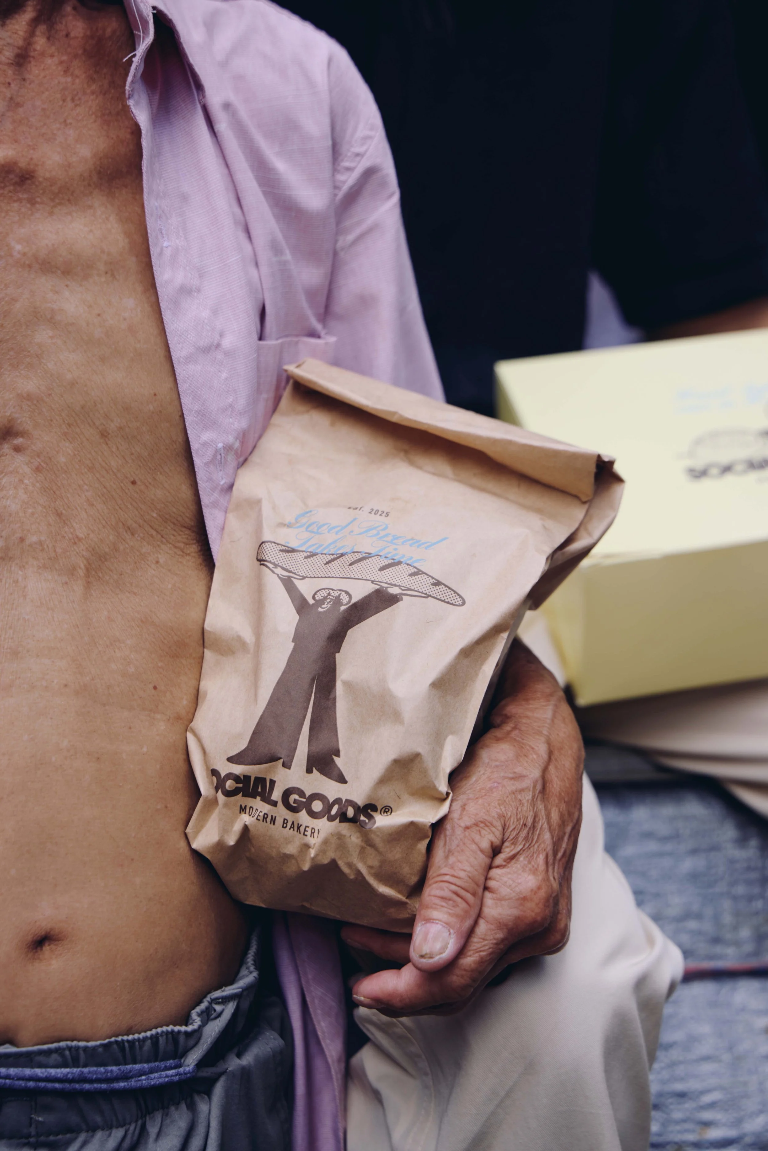 An elderly hand holds a paper bag from Social Goods bakery against the textures of a traditional Hong Kong street.