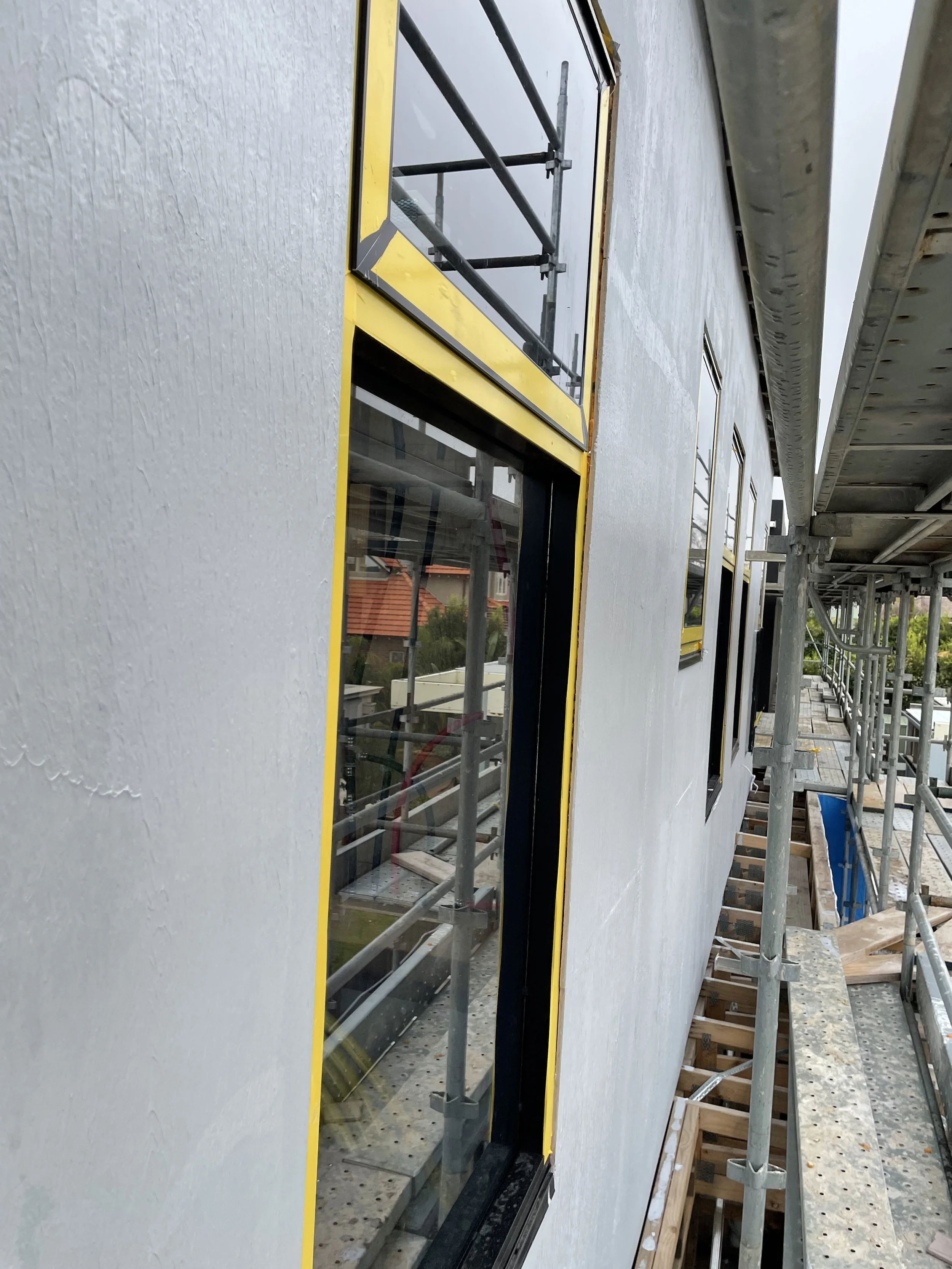 Exterior wall of a building under construction, with multiple windows framed with yellow, and scaffolding surrounding the structure.