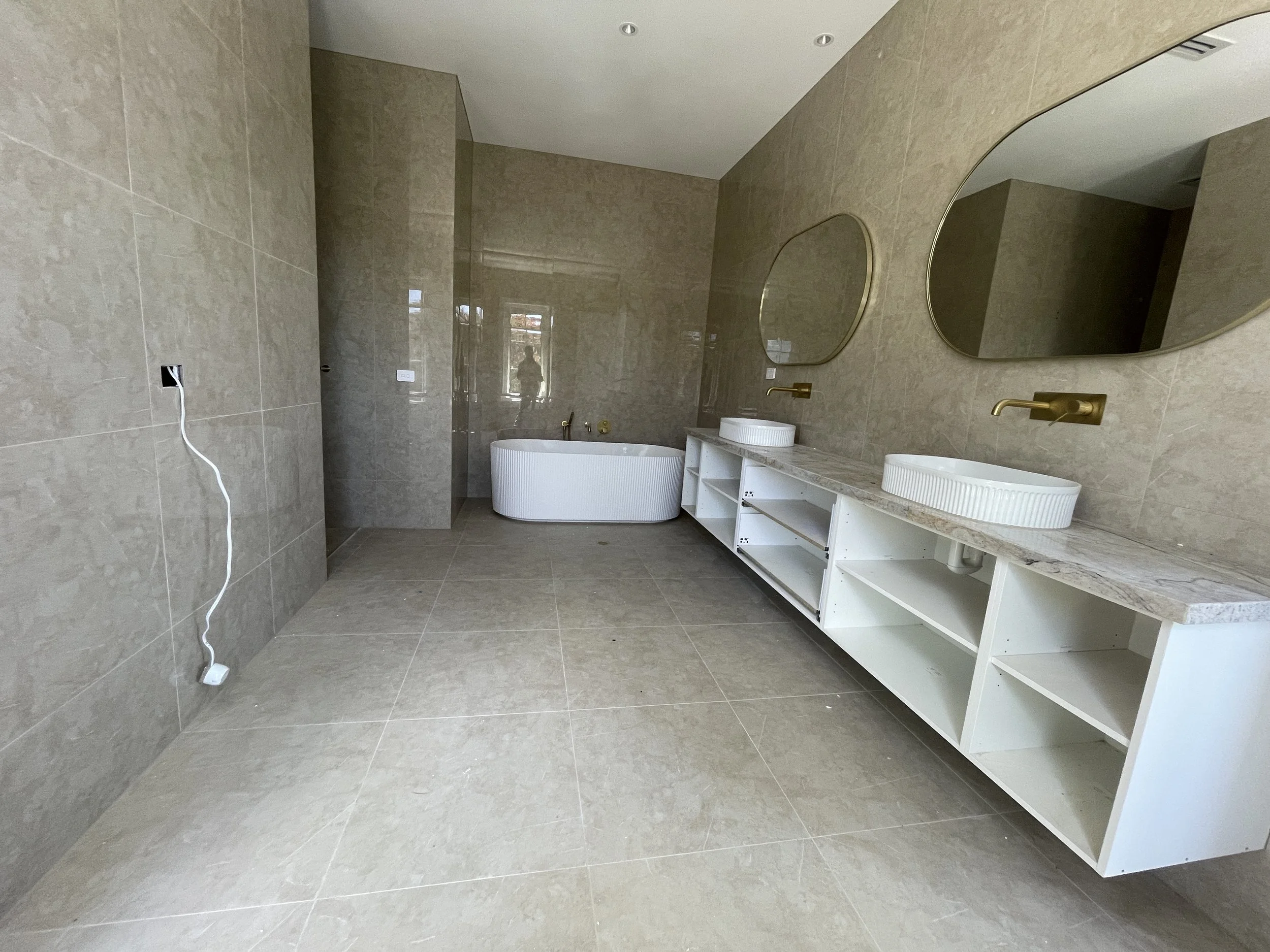Bathroom with beige tiled walls and floor, two round mirrors above white marble countertop, gold fixtures, two vessel sinks, and a freestanding bathtub at the back. An electrical outlet with a white cord hanging, visible on the left wall.