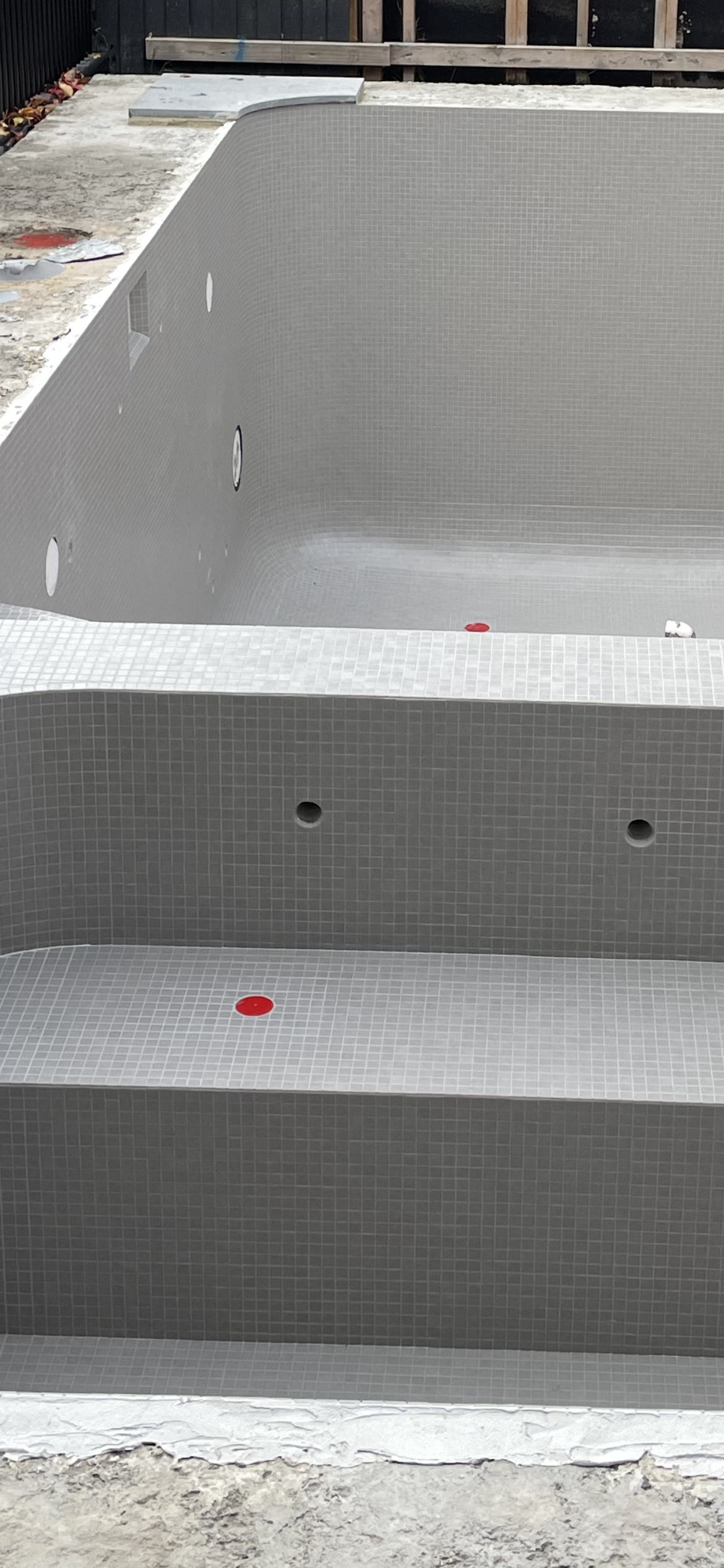 Empty swimming pool with gray tiles and red and white caps inside, under construction, with concrete and construction materials around.