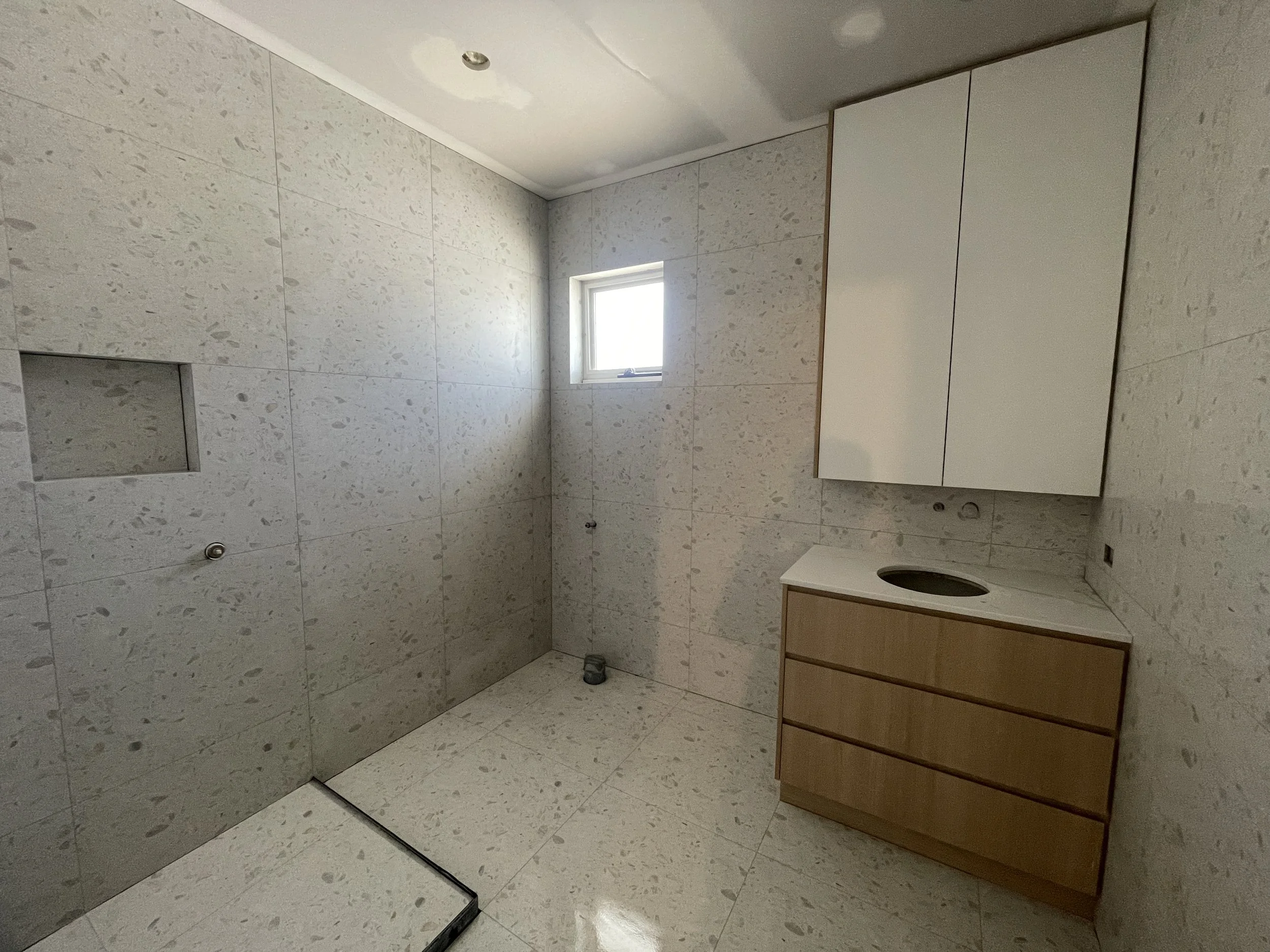 Empty bathroom with beige tiled walls and floor, a small window, a white wall cabinet, and a wooden vanity with a sink cutout.