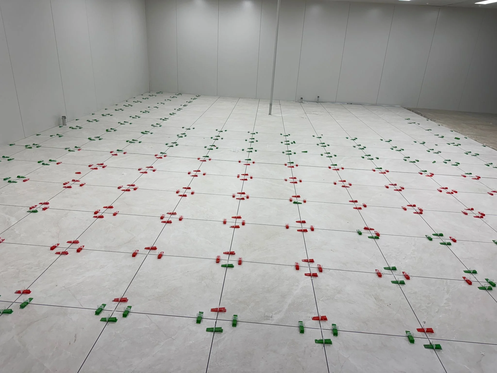 A room with tiled floor and empty walls, with green and red plastic spacers arranged in grid patterns on the floor, used for tiling installation.