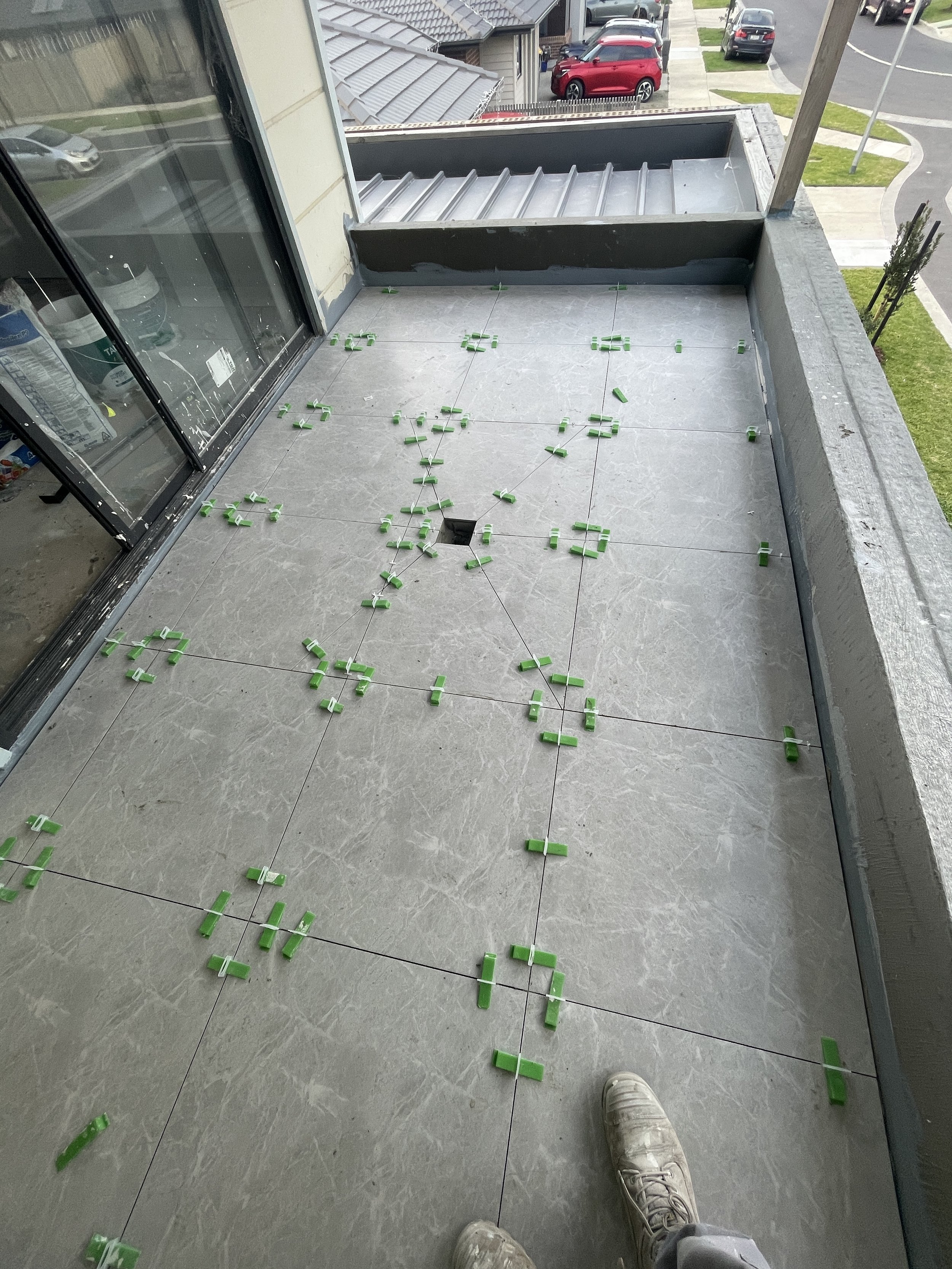 Balcony floor with green tile spacers and grout lines, partially installed tiles, and a person's white shoes visible at the bottom.