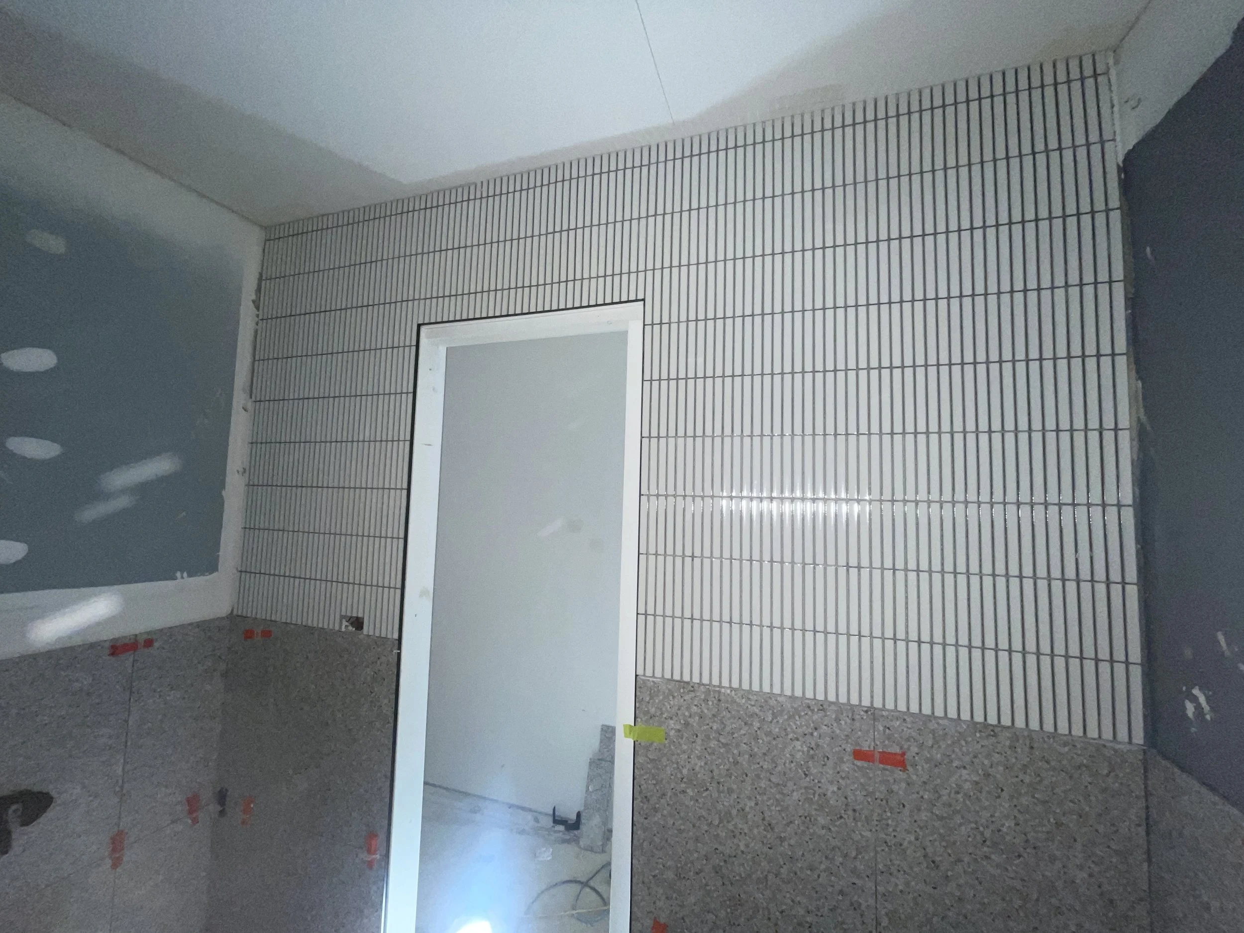 Bathroom wall partially tiled with small white rectangular tiles, with a doorway leading to another room, and visible construction materials and tape on the wall.