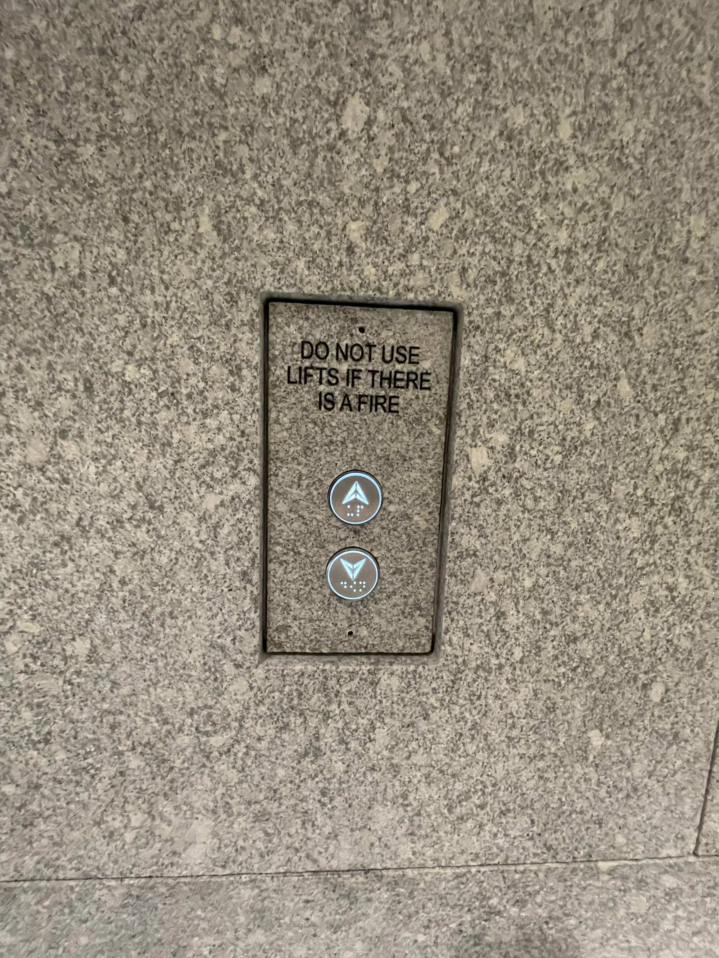 Elevator control panel with two illuminated buttons, one for going up and one for going down, mounted on a textured wall. A sign above states, 'DO NOT USE LIFTS IF THERE IS A FIRE'.