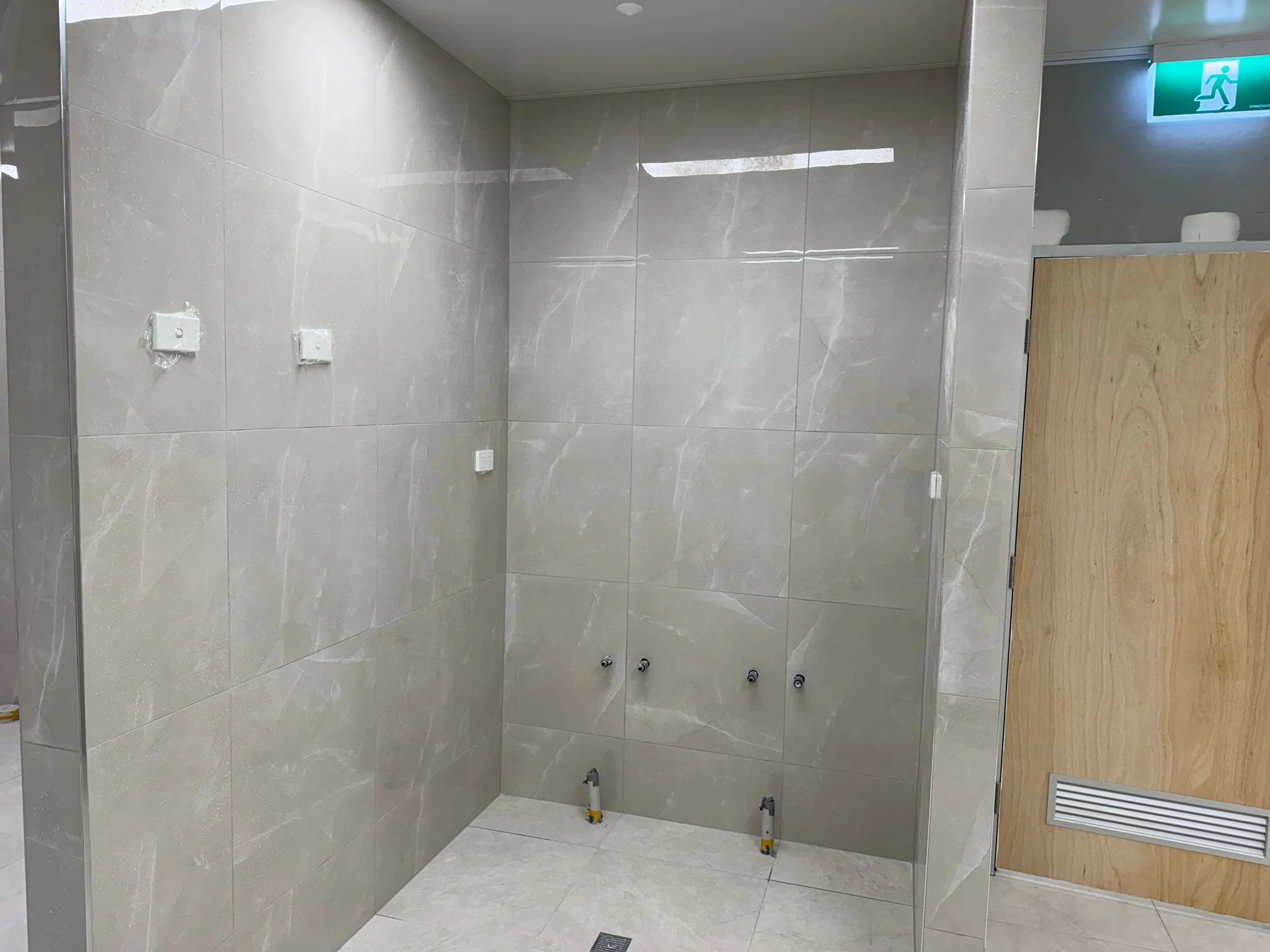 Empty bathroom or washroom with tiled walls and floor, several plumbing fixtures protruding from the wall, and a wooden door with a vent, with an emergency exit sign above.