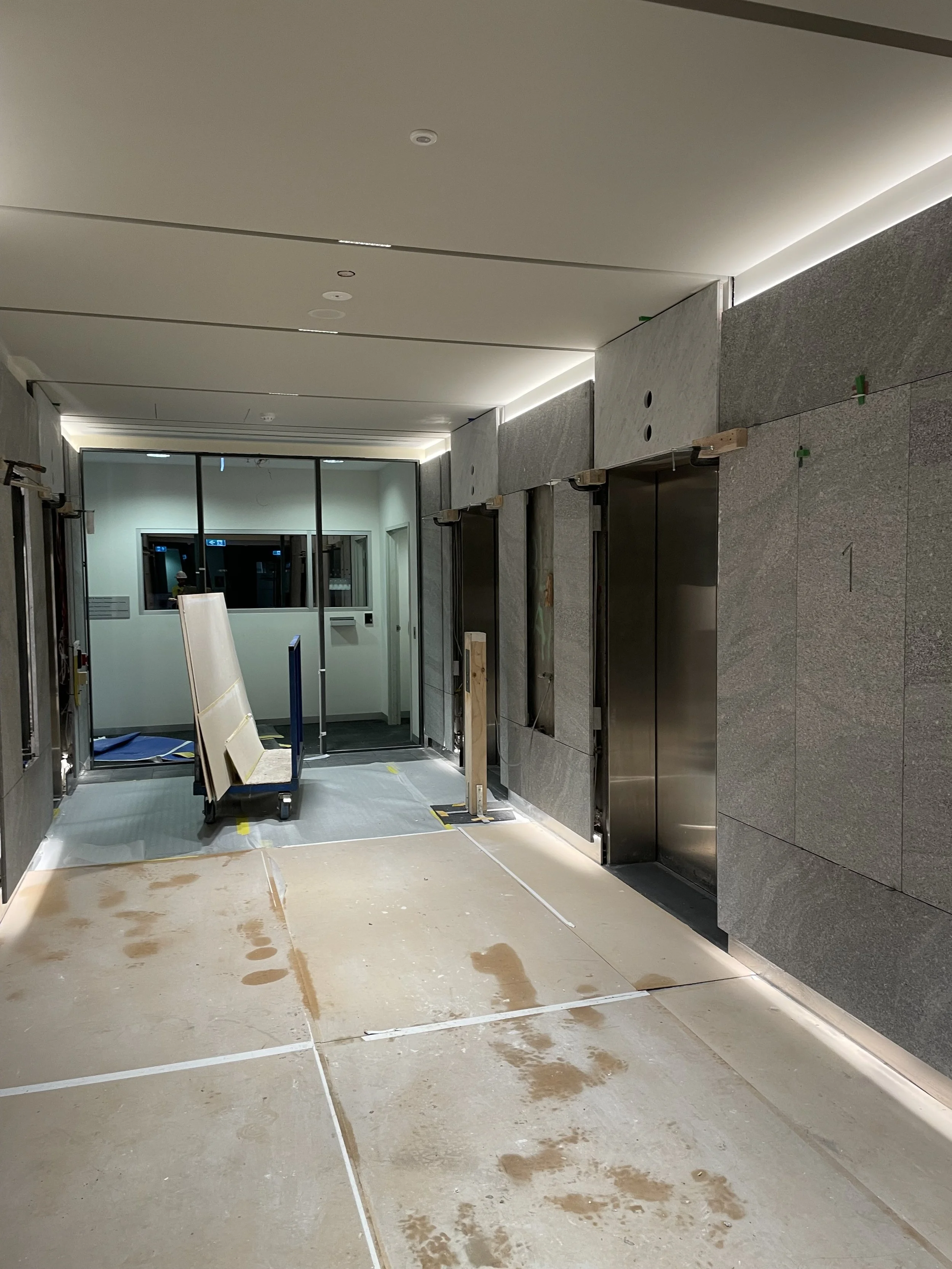 Construction site of an elevator lobby with untiled flooring, ongoing installation of wall panels, and elevator doors. There are building materials and equipment in the space.