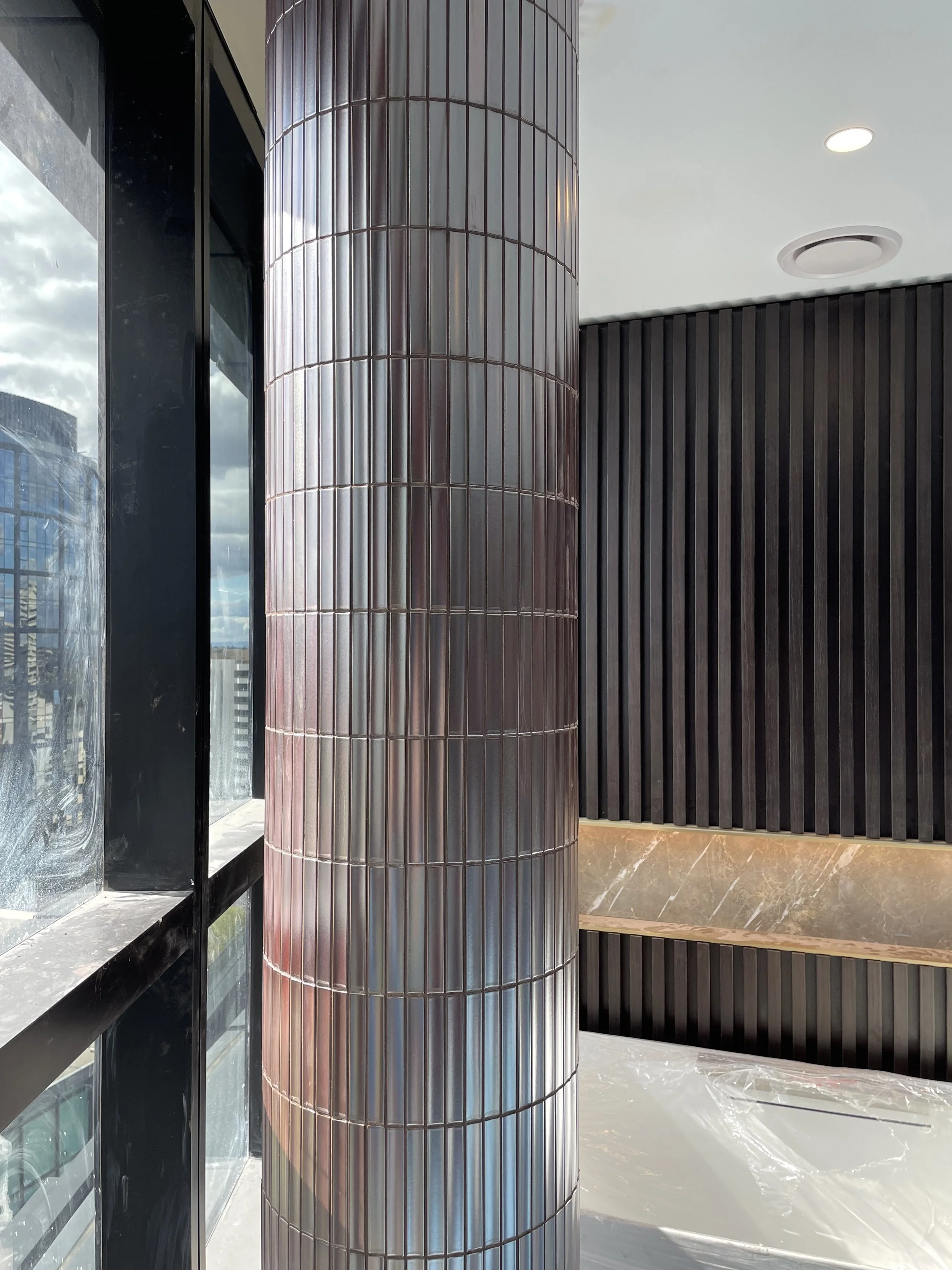 Modern interior with a large cylindrical metallic pillar, black textured wall with vertical grooves, marble bench, and a window with city view and cloudy sky.
