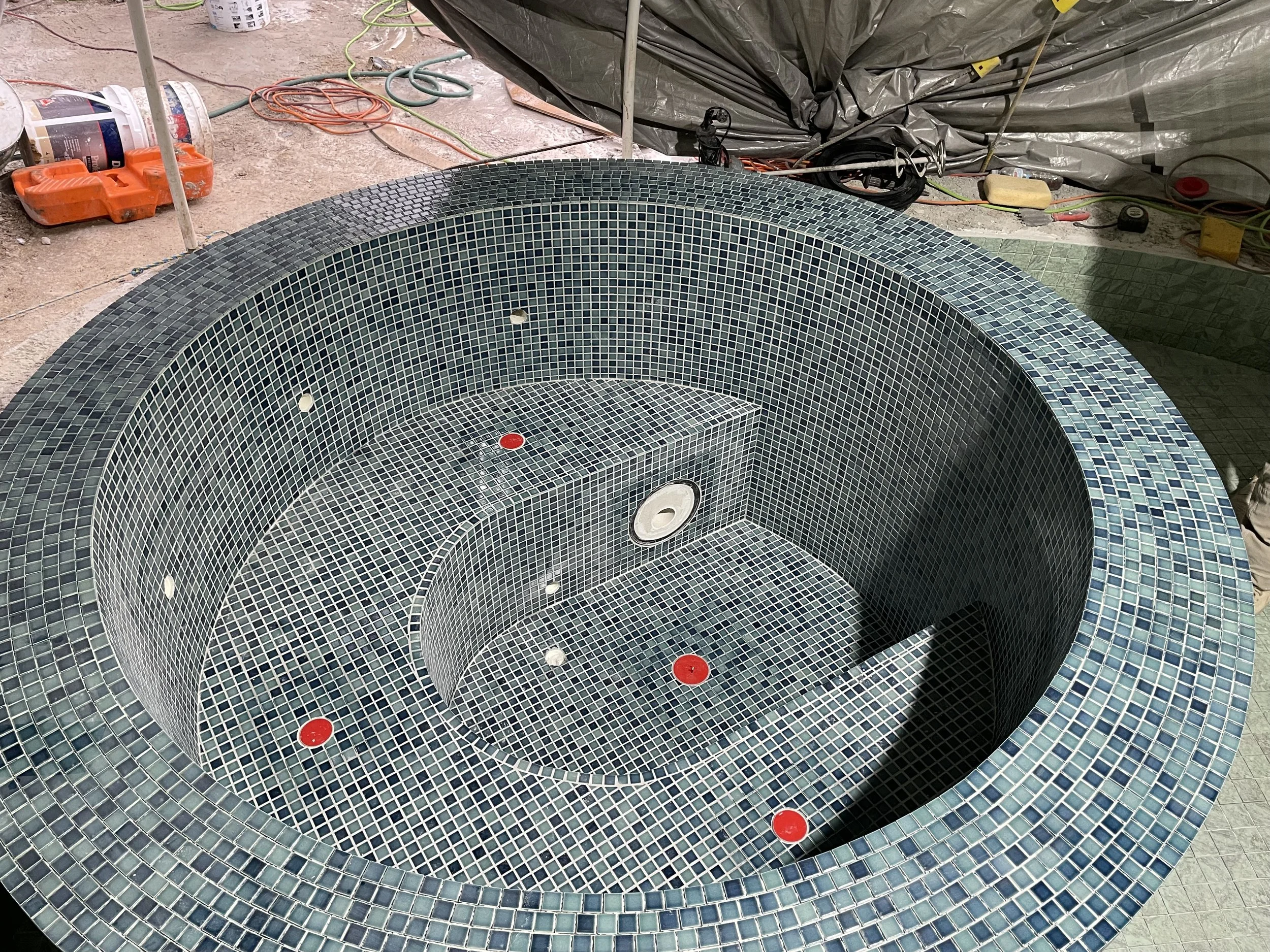Large circular mosaic hot tub under construction with small square tiles in shades of blue and black, featuring built-in seating and jet access, surrounded by construction tools and supplies.