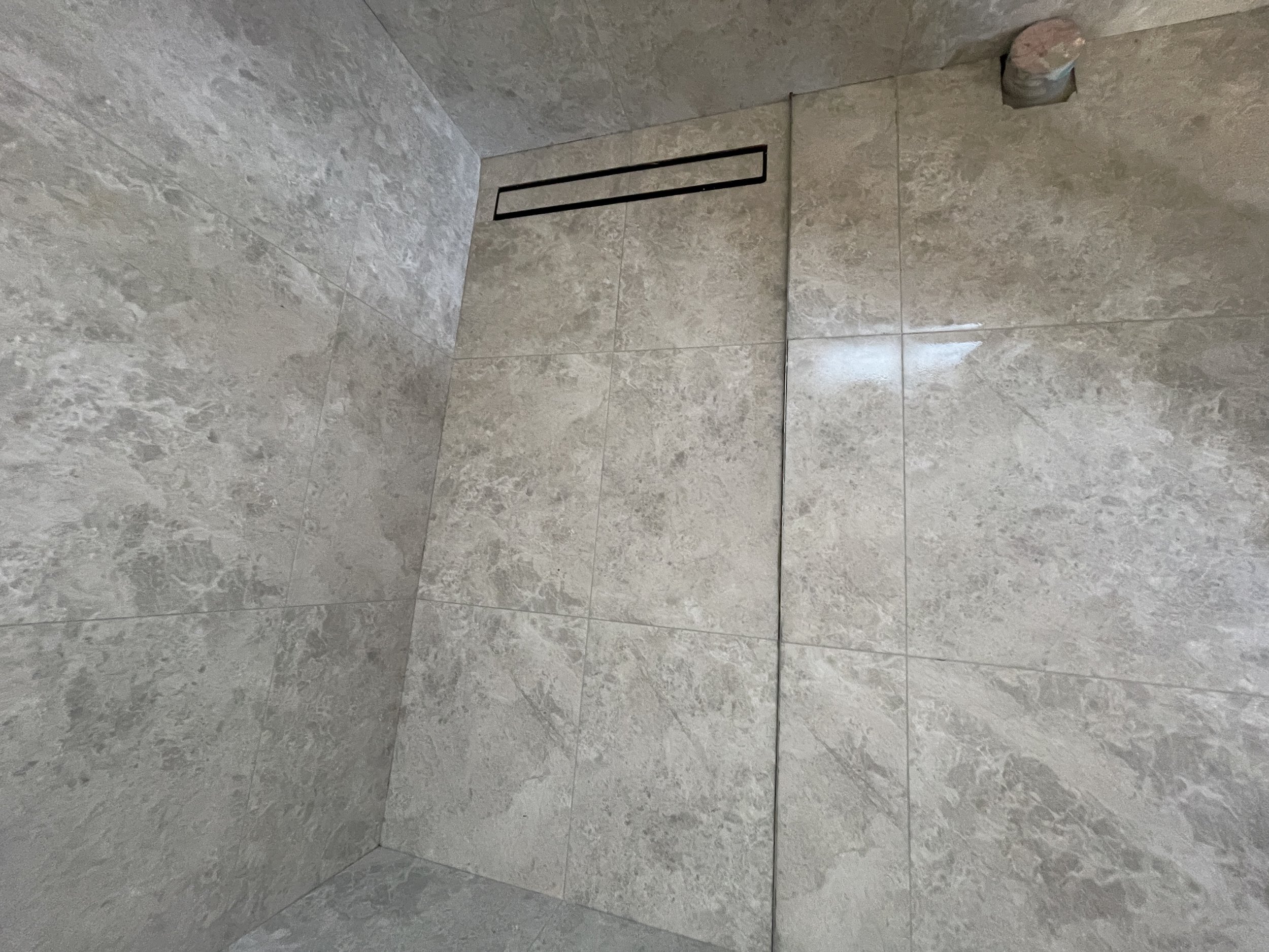 Interior of a shower with large beige marble tiles on the walls and floor, a rectangular black drain, and a pipe protruding from the ceiling.