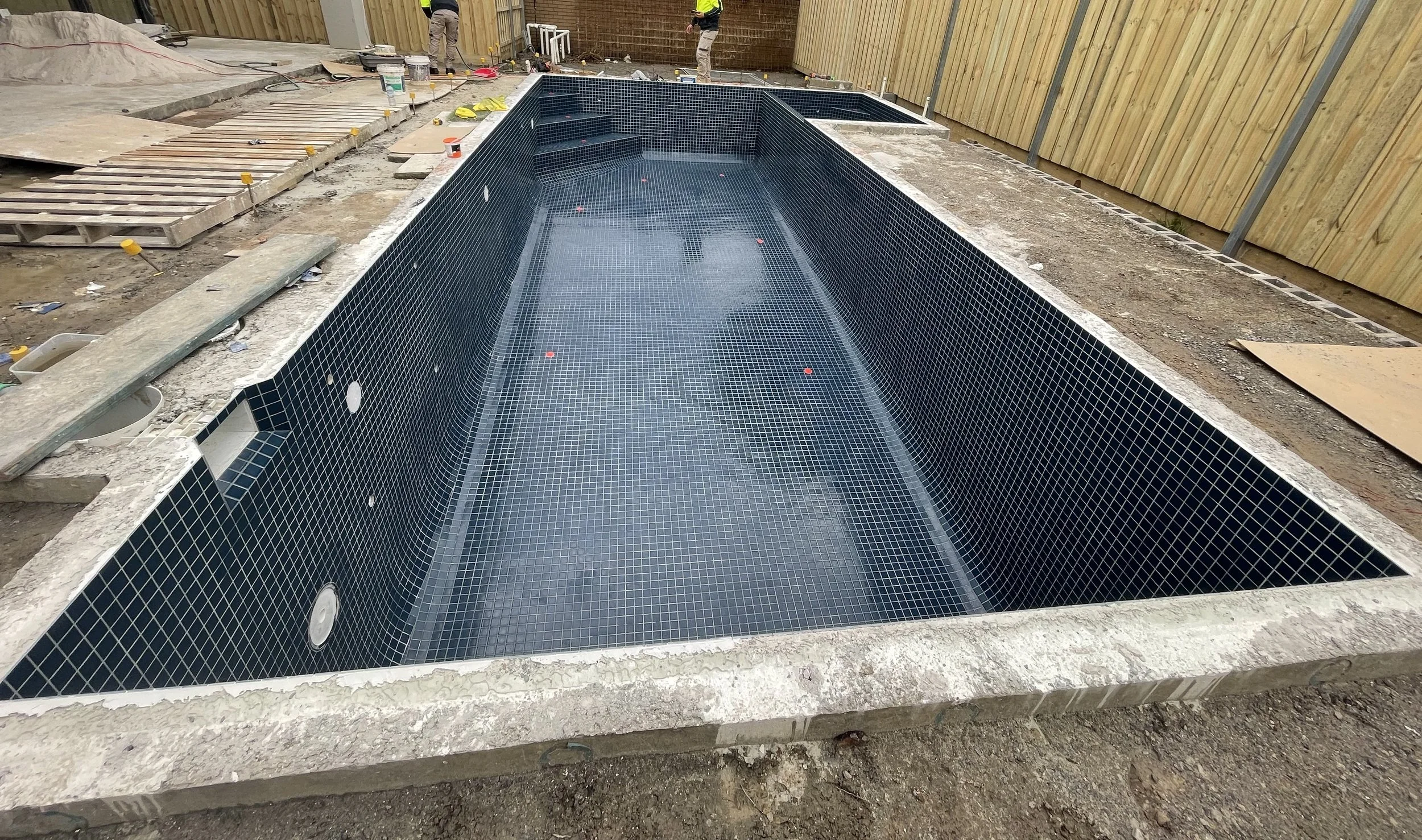 Under construction in-ground swimming pool with black tile interior, surrounded by a concrete deck and a partially fenced backyard.