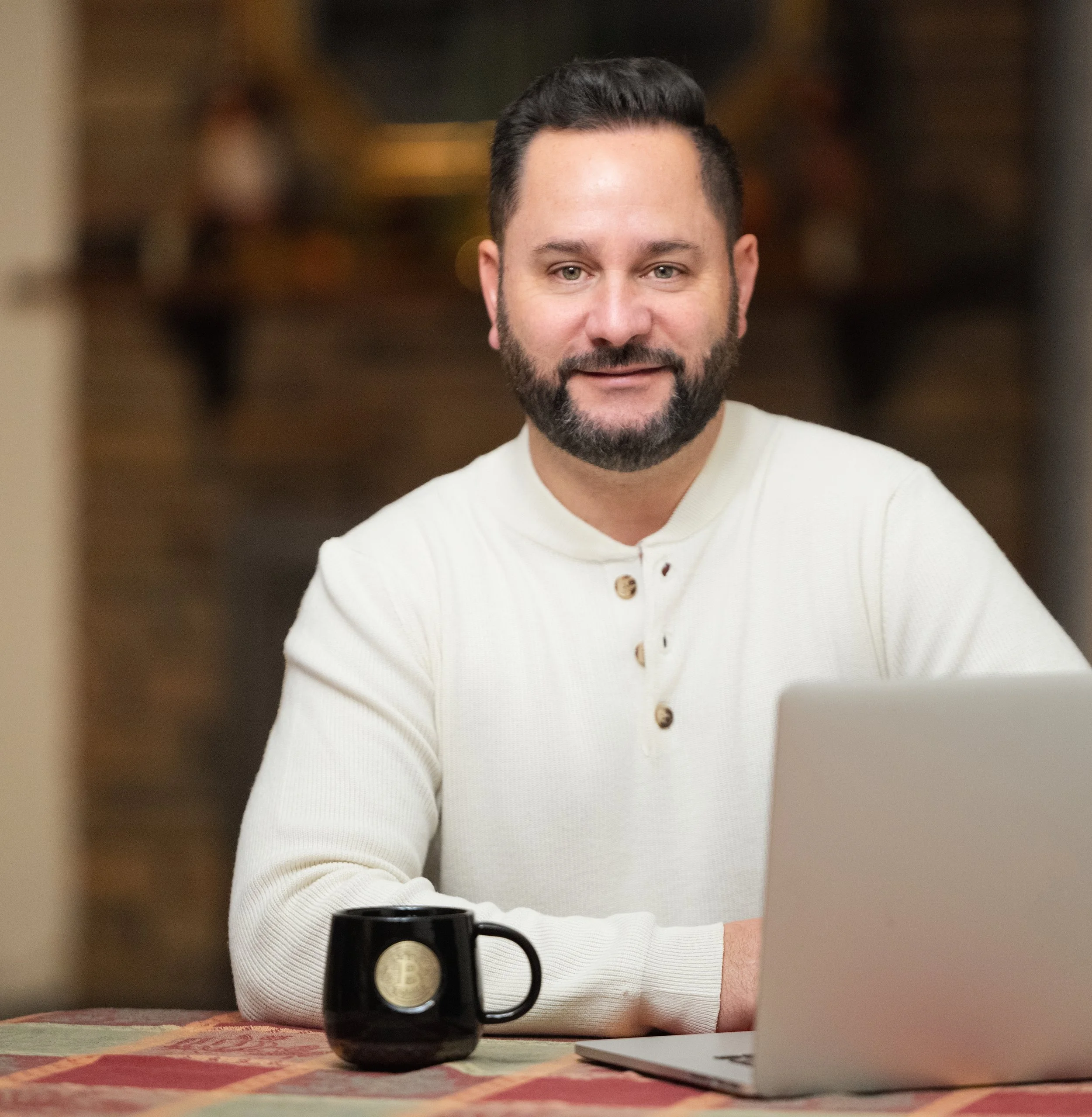 A man with dark hair and a beard sitting at a table with a laptop and a black mug with a Bitcoin symbol, smiling at the camera in a warmly lit room.