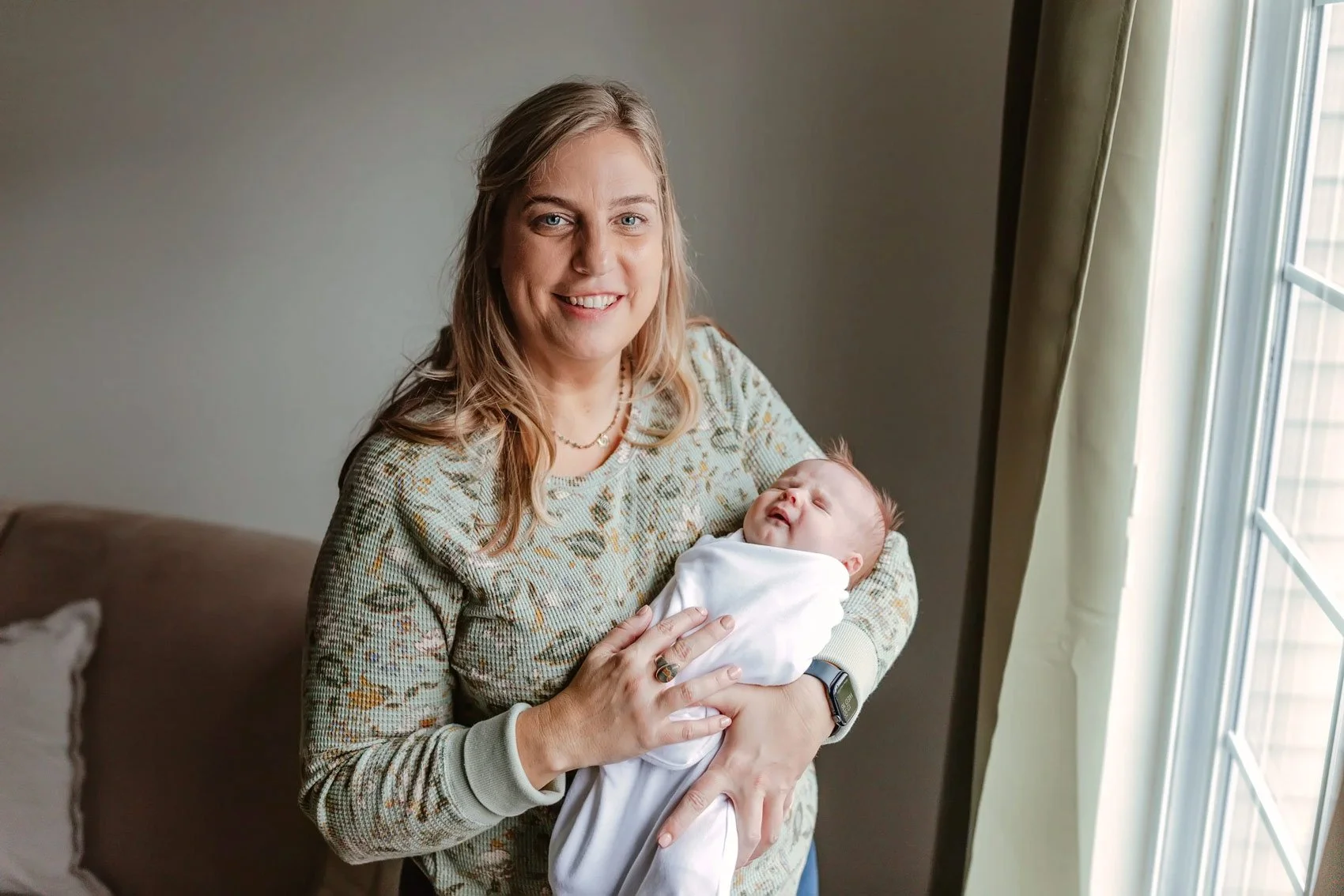 A smiling woman holding a newborn baby wrapped in a white blanket near a window.