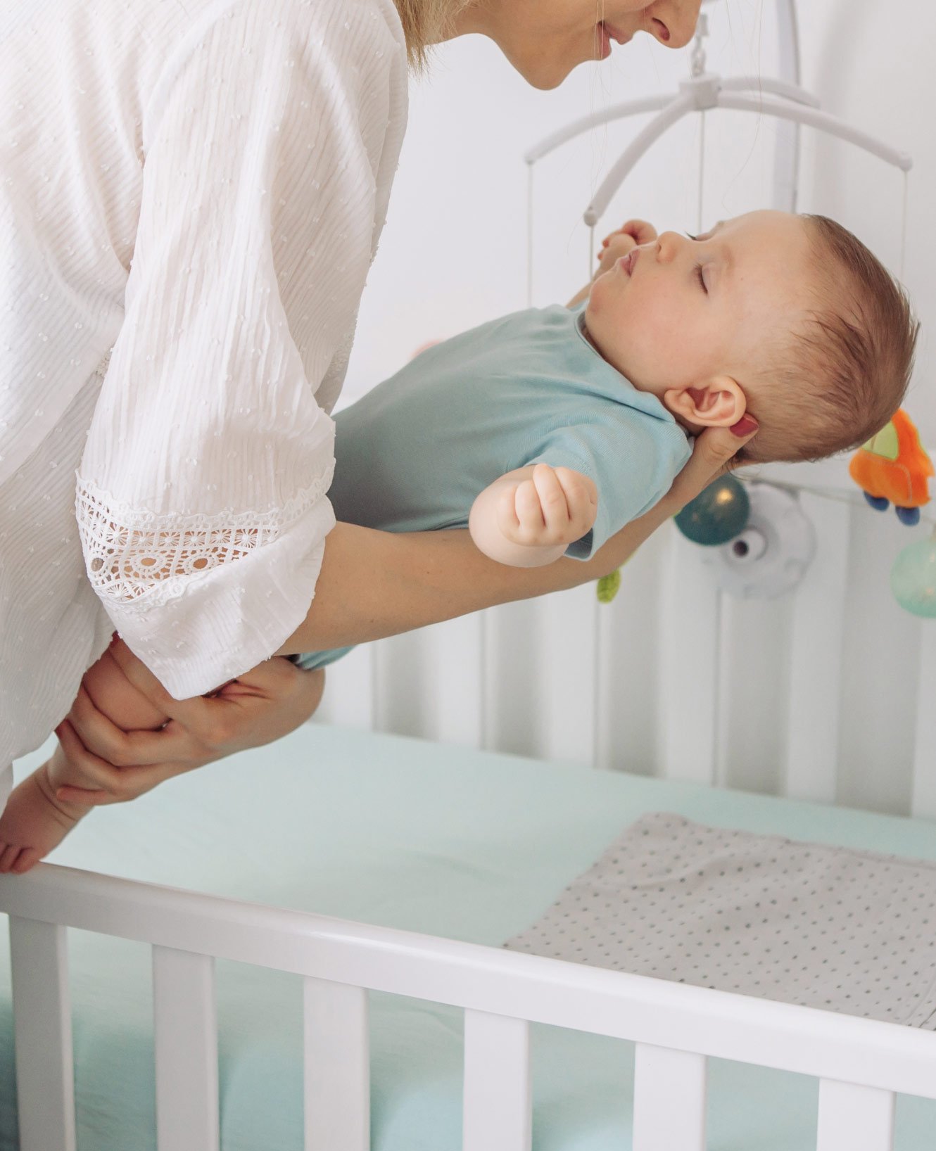 A woman holding a sleeping baby in a nursery. The baby is dressed in a blue outfit, and the woman is wearing a white blouse with lace detail on the sleeve. There are colorful hanging toys on the crib in the background.