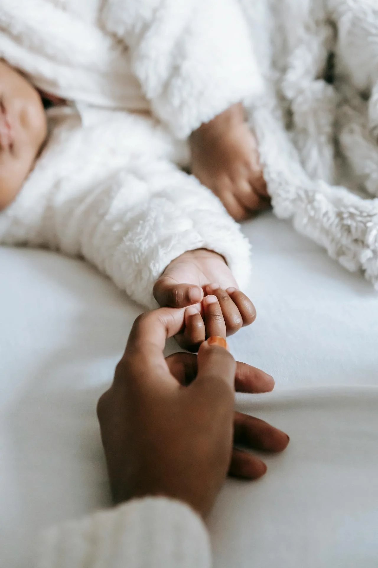 A person's hand holding a baby's hand, with a baby lying on a bed wearing a white, fluffy outfit.
