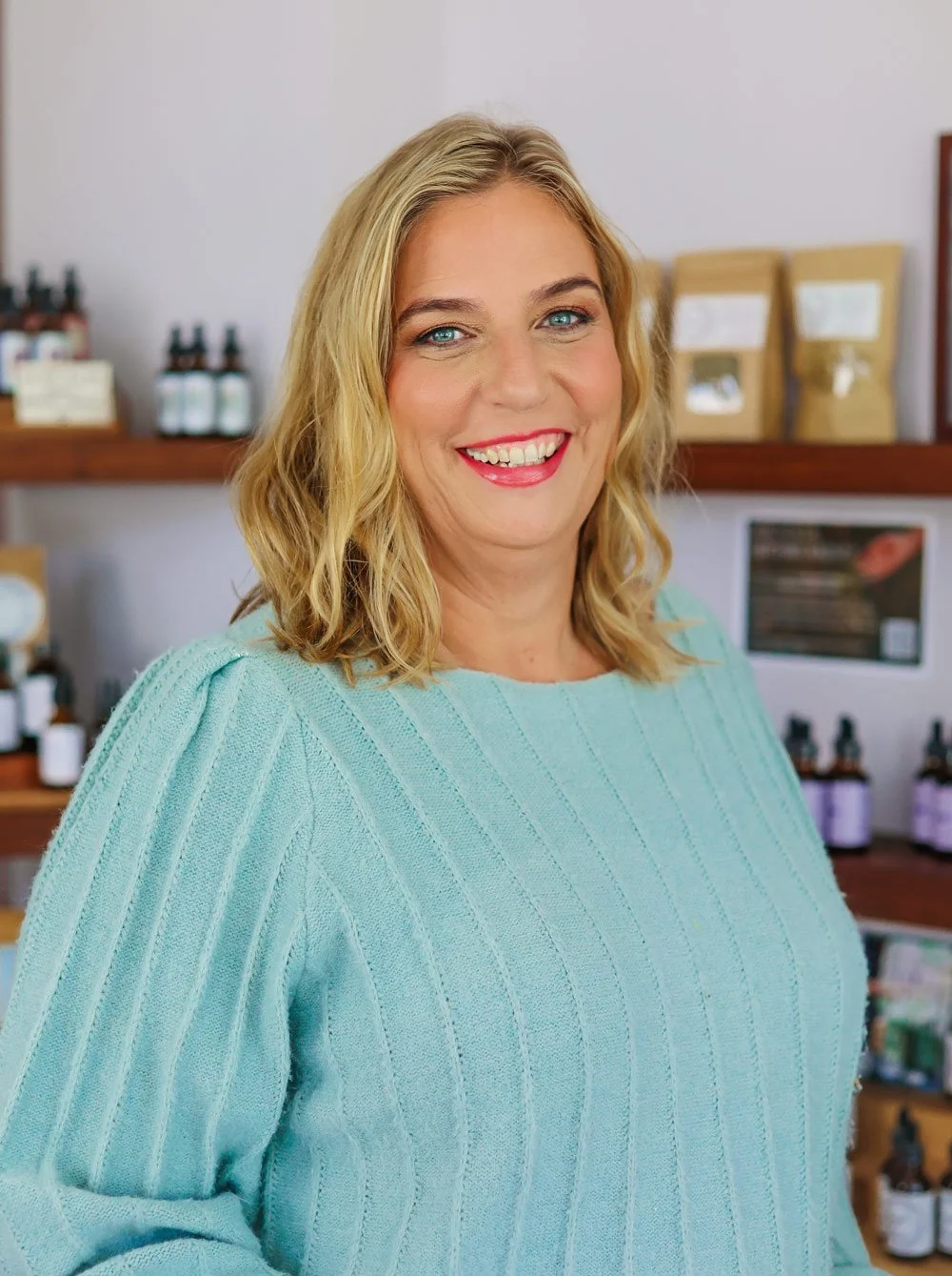 Smile woman with blonde wavy hair wearing a light blue sweater inside a shop with shelves of bottles and packages in the background.