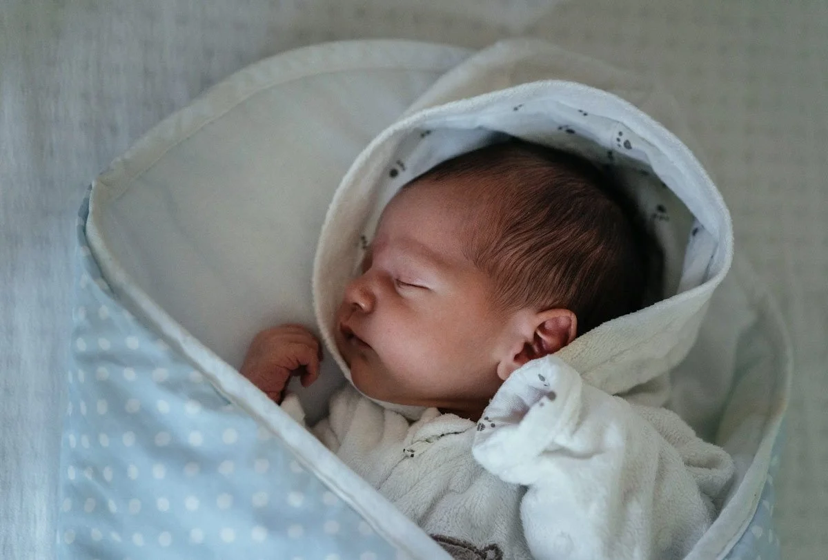 A sleeping newborn baby wrapped in white clothing and lying in a blue polka-dotted bassinet with a hood.