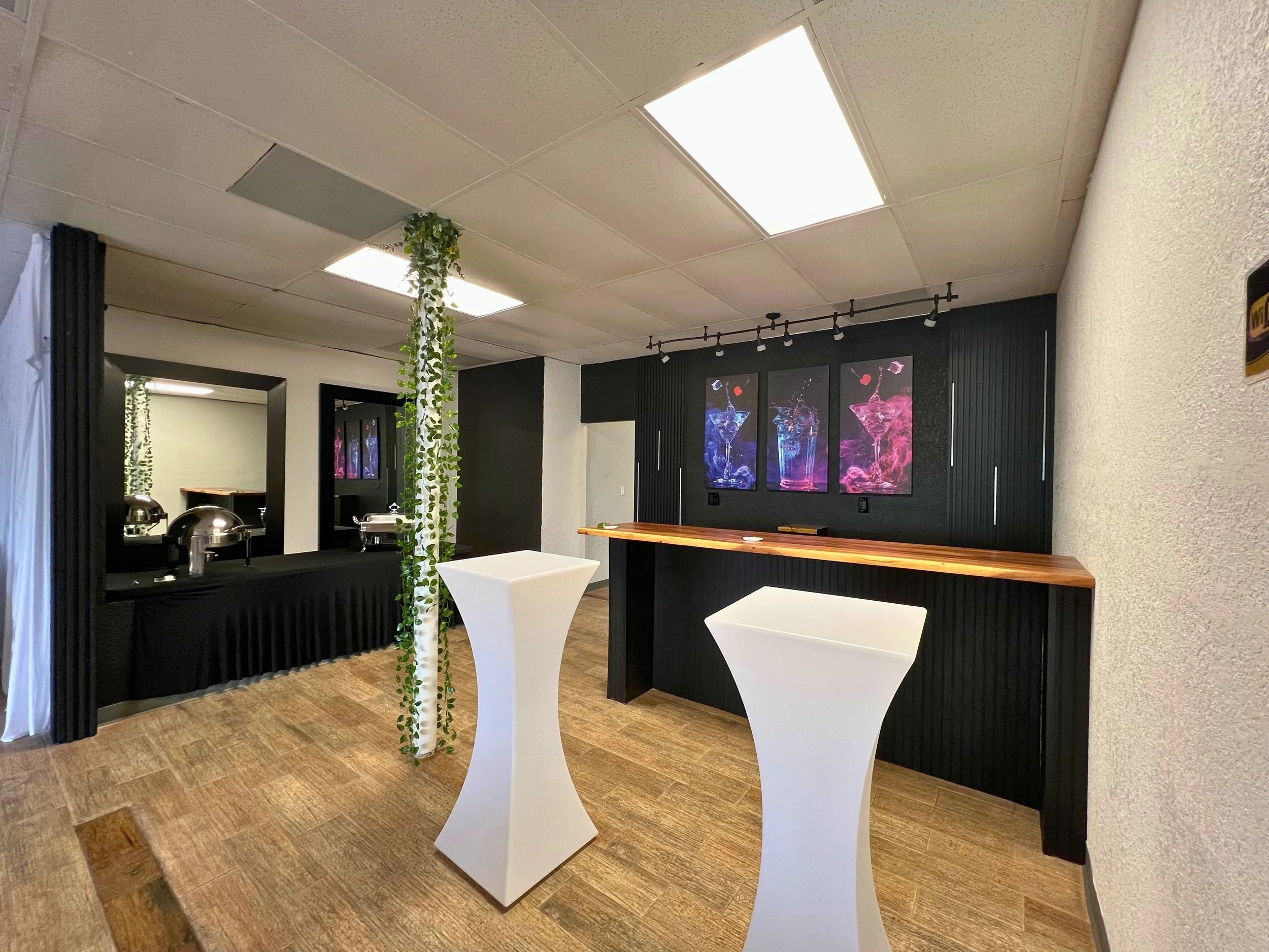Event space with black and white decor, wood flooring, two tall white cocktail tables, a black buffet table with chafing dishes, and framed colorful abstract artwork on the black wall.