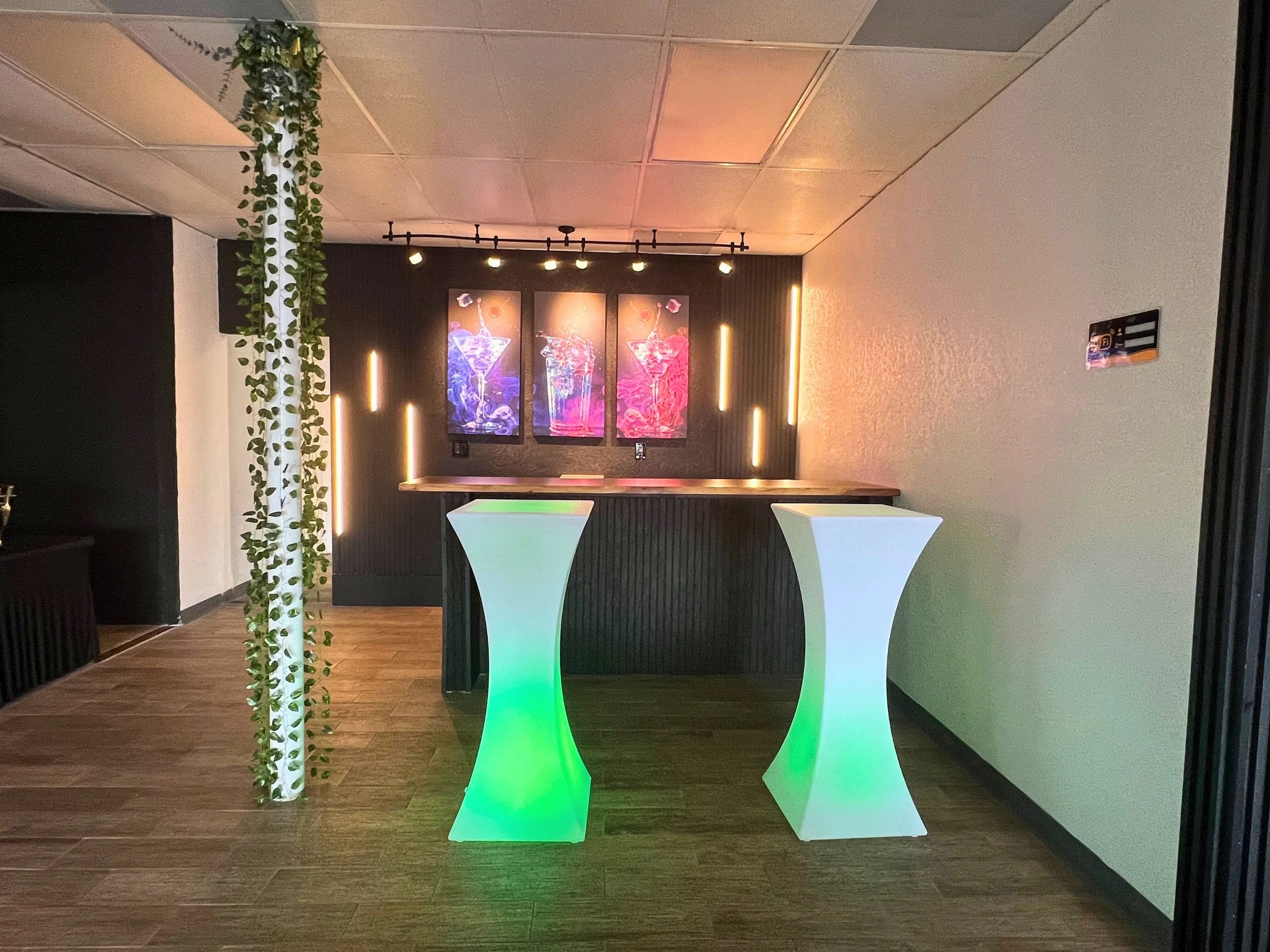 A modern bar area with two illuminated cocktail tables in green and white, dark wood flooring, black and white walls, and three colorful artwork prints of cocktails on the back wall, accented by vertical light strips.