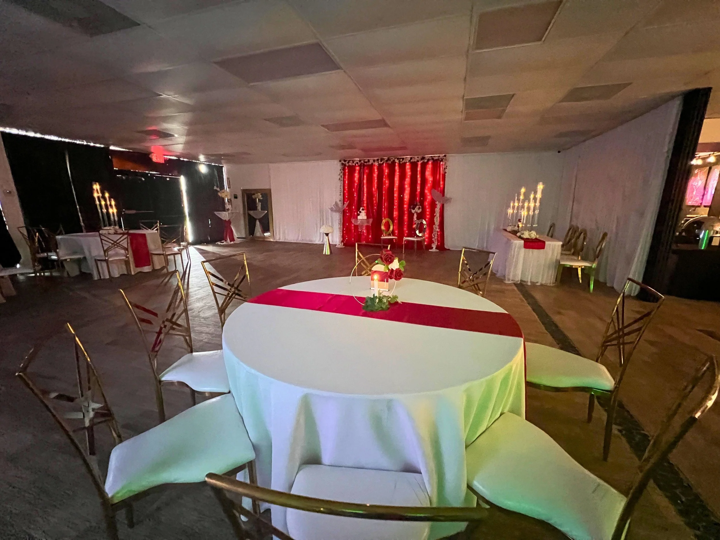 Decorated event space with white and red themed decorations, round table with white tablecloth and red runner, chairs arranged around the table, backdrop with red curtain, white flowers, and number '05' in the background, some tables with white candl