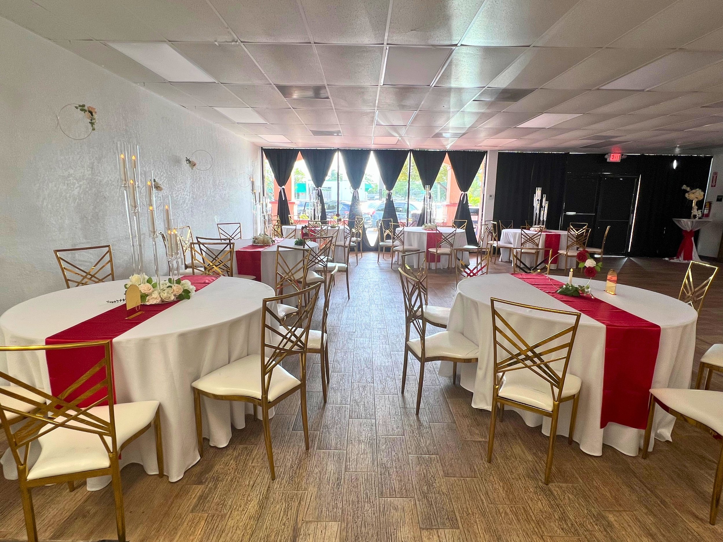 Party or event venue decorated with white round tables and gold chairs, featuring red table runners, floral centerpieces, and tall candle holders with candles. Large windows with black curtains at the back let in natural light.