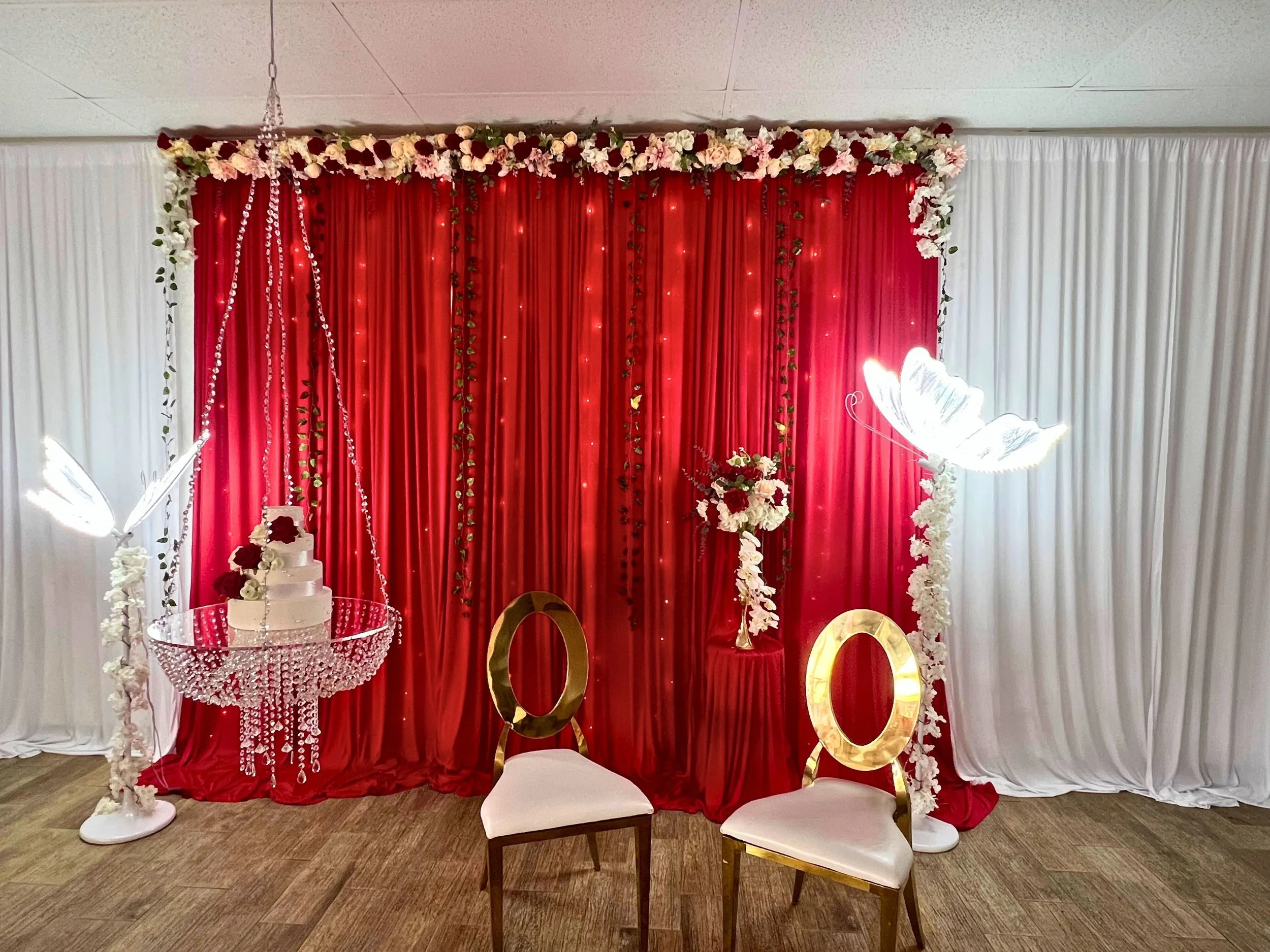 Decorative wedding backdrop with red and white drapes, floral arrangements, butterfly light fixtures, a cake, and two chairs.