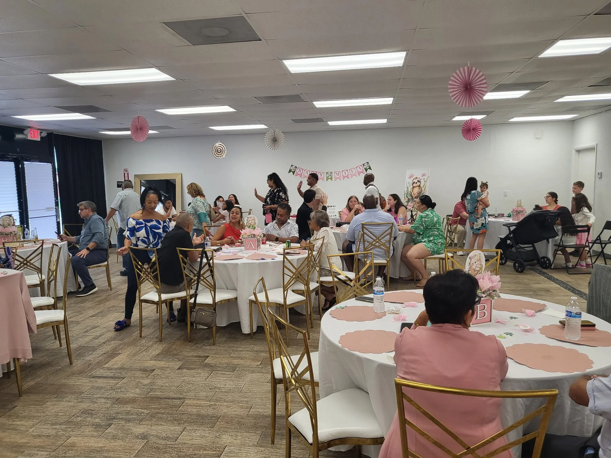 A decorated indoor party room with round tables, some with pink and white decorations, and guests seated and standing, celebrating a baby shower. Pink and white paper decorations hang from the ceiling, and a banner with the words 'Baby Moon' is on the wall.