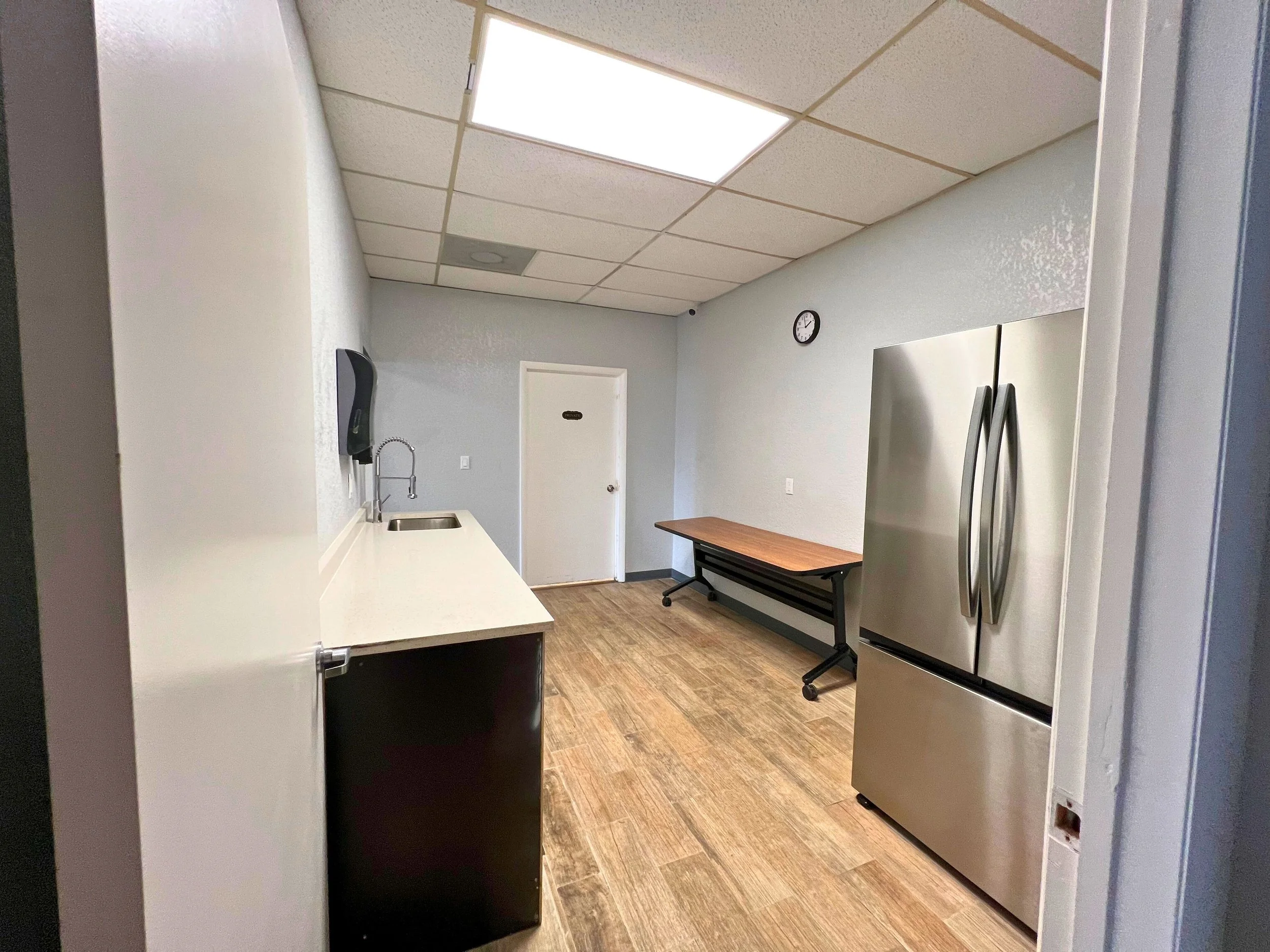 Empty break room with a small sink and paper towel dispenser on the left, a stainless steel refrigerator on the right, a folding table along the back wall, light-colored walls, a wood laminate floor, a clock on the wall, and ceiling tiles with fluore