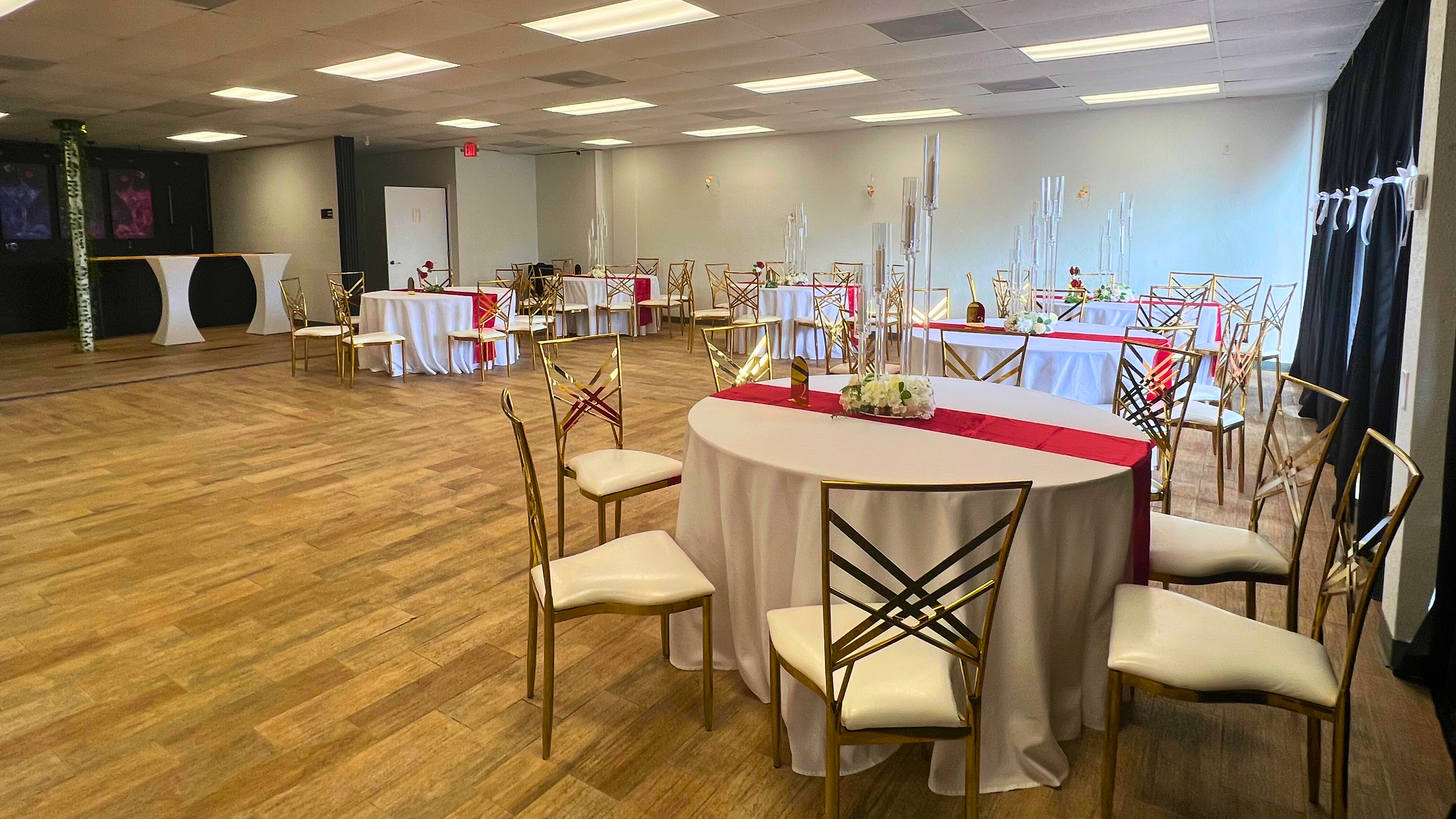 Banquet hall decorated for a celebration with round tables covered in white tablecloths, red table runners, floral centerpieces, and gold chairs, with a wooden floor and a ceiling with overhead lights.