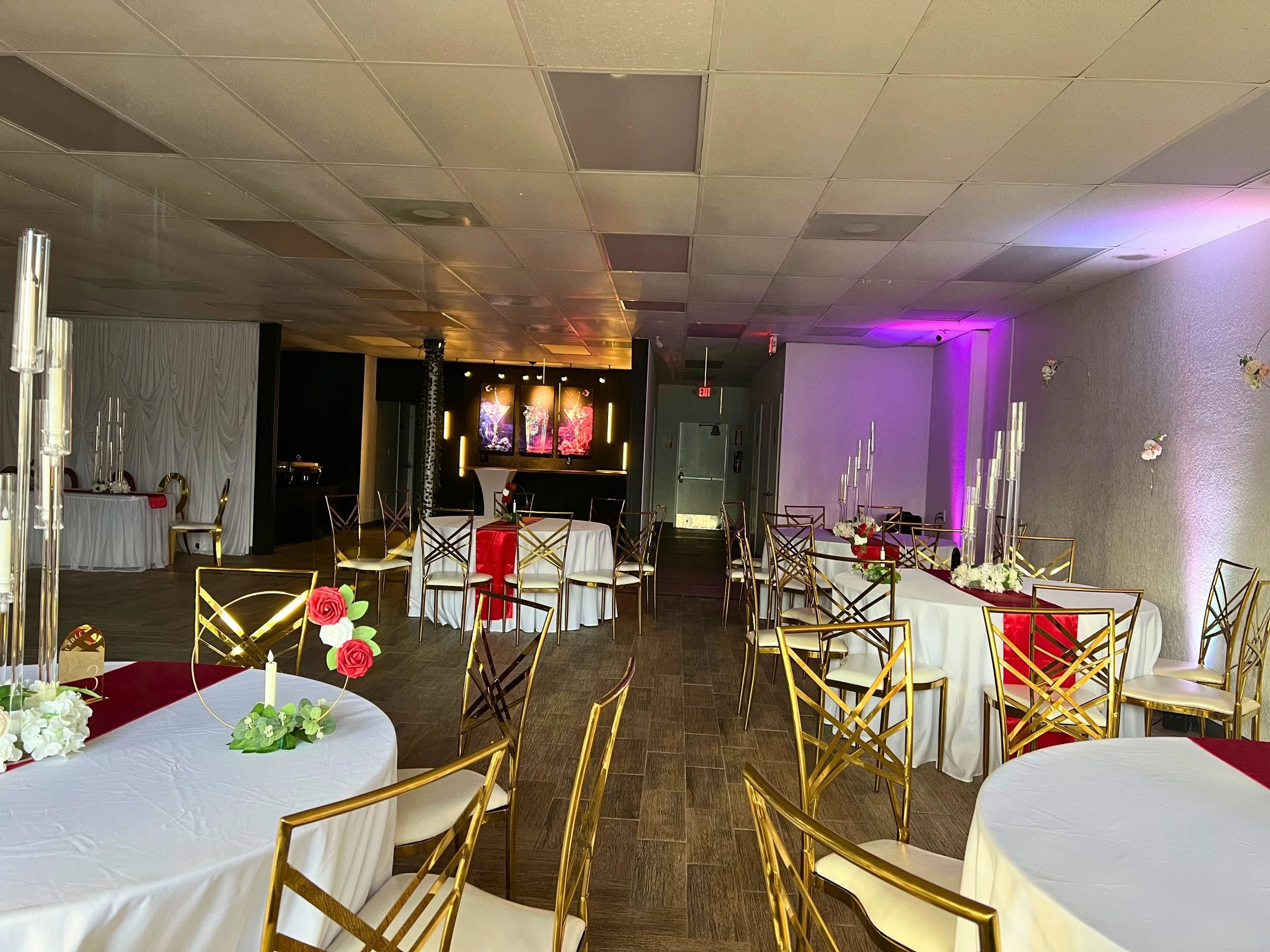 Decorated banquet hall with round tables covered in white tablecloths and gold chairs, floral centerpieces, tall white candles, and purple lighting accents.
