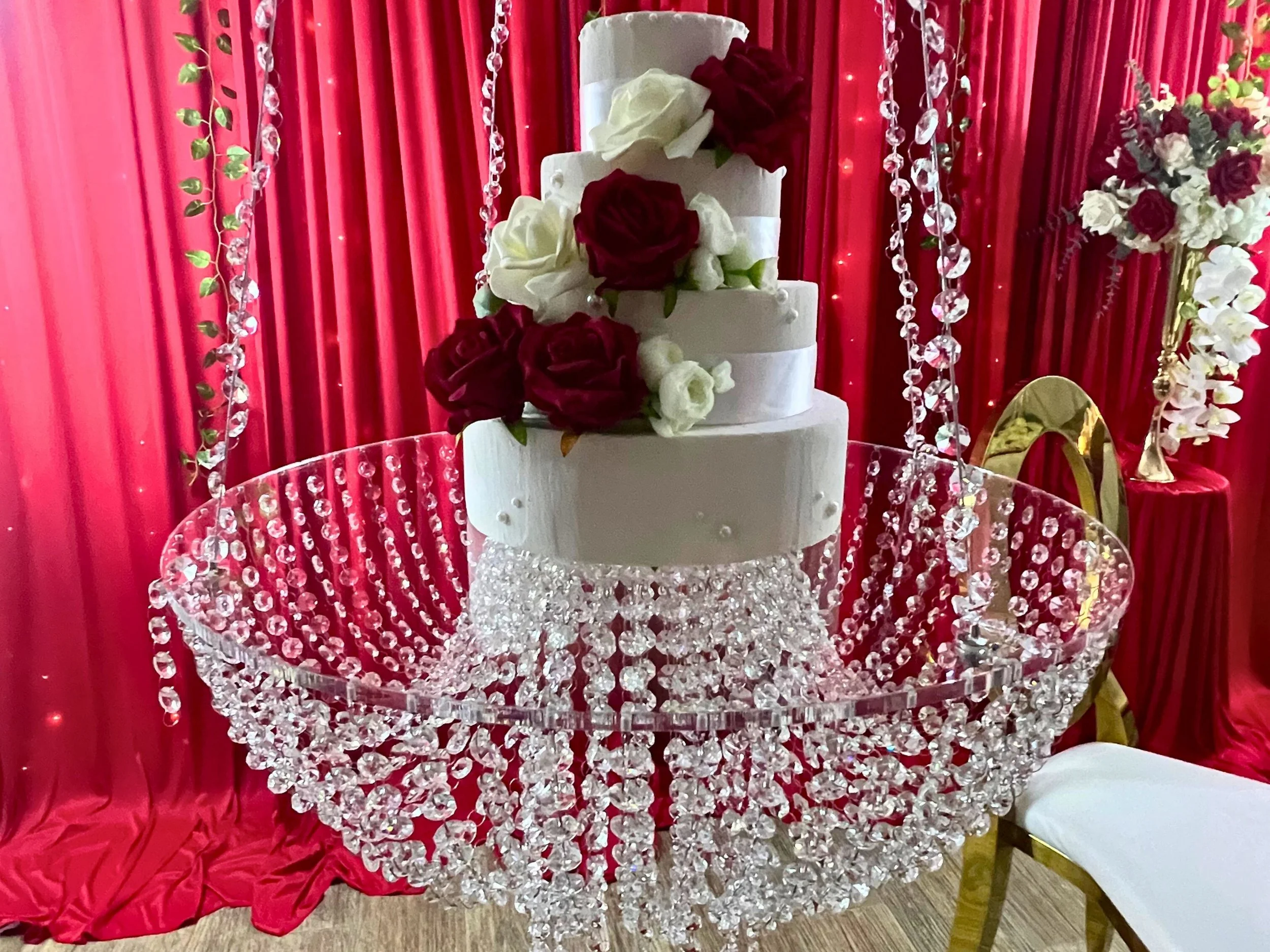 A multi-tiered white wedding cake with red and white roses on top, displayed on a large crystal chandelier table with a red curtain backdrop decorated with hanging crystal strands and floral arrangements.