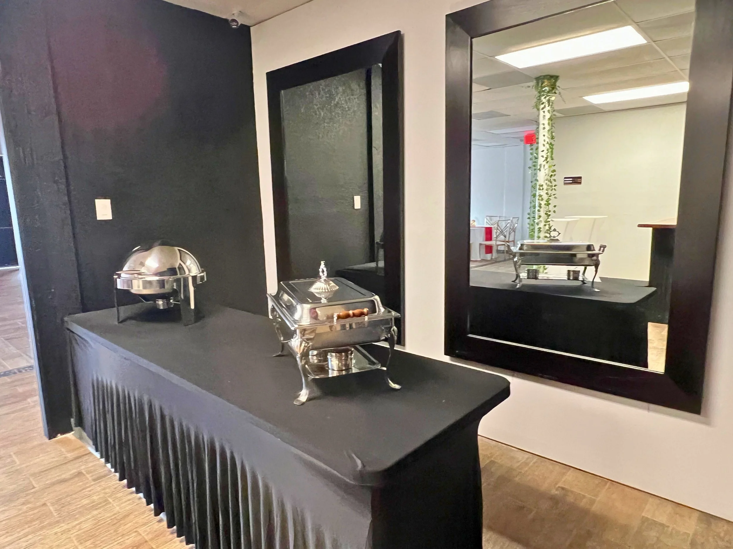 Buffet table with covered chafing dishes and a mirror reflecting a seating area with a decorative plant in the background.