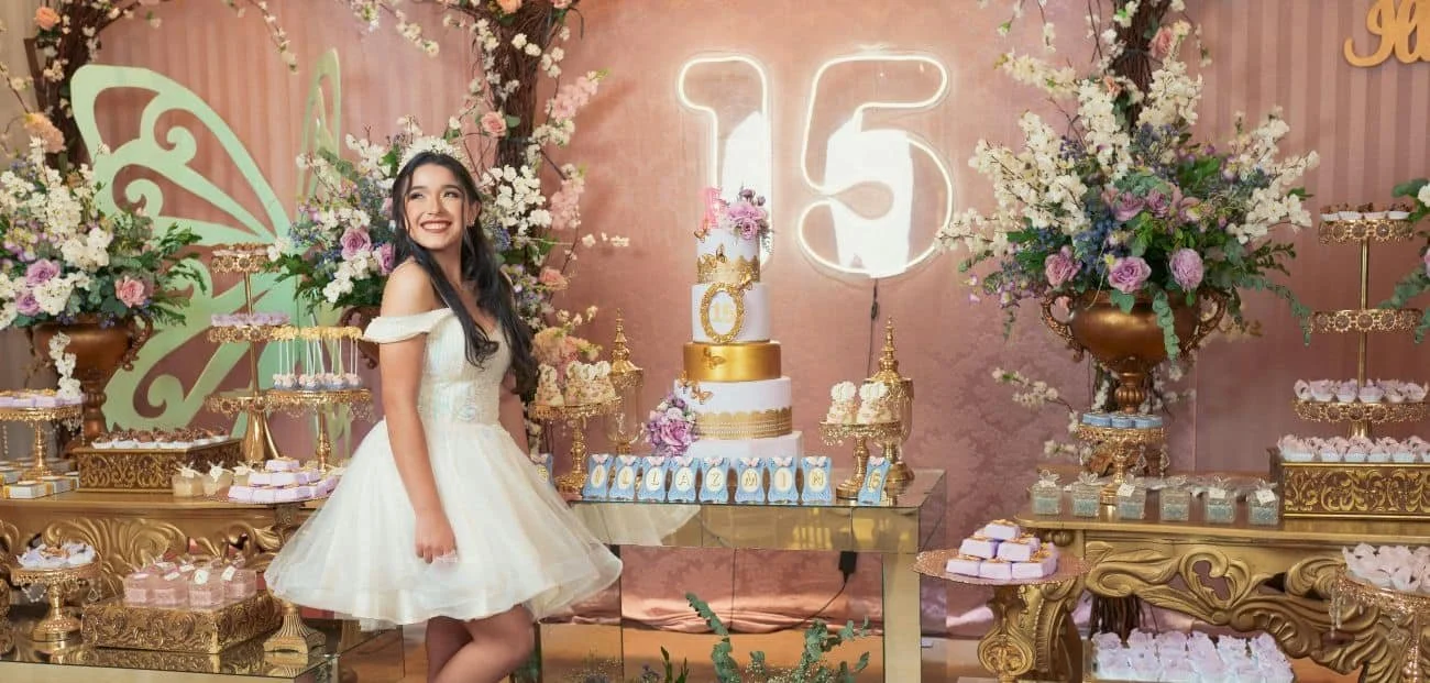 A young woman in a white dress stands in front of a decorated table with a birthday cake and pink and purple flowers. There are large floral arrangements and a neon number 15 in the background, indicating a 15th birthday celebration.