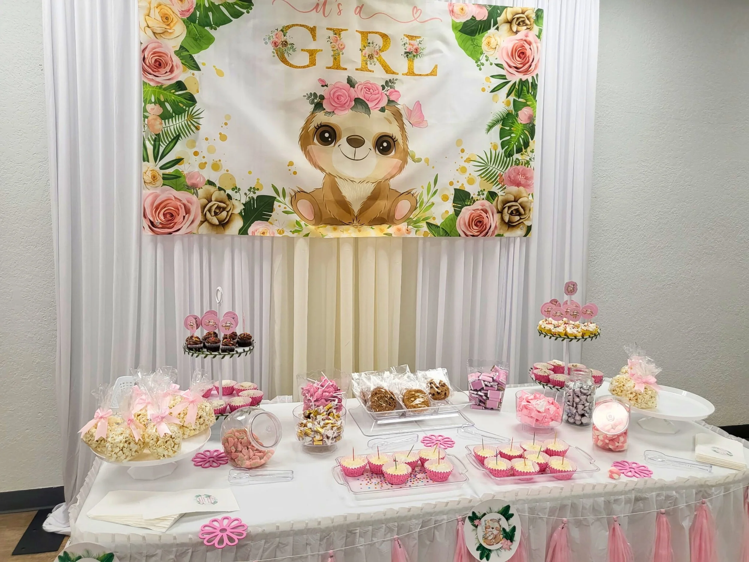 Baby girl-themed party table with pink and white treats, including cupcakes, popcorn, marshmallows, and cookies, decorated with pink bows and flower cutouts, with a large banner featuring a cartoon sloth and floral designs, reading 'It's a Girl'.
