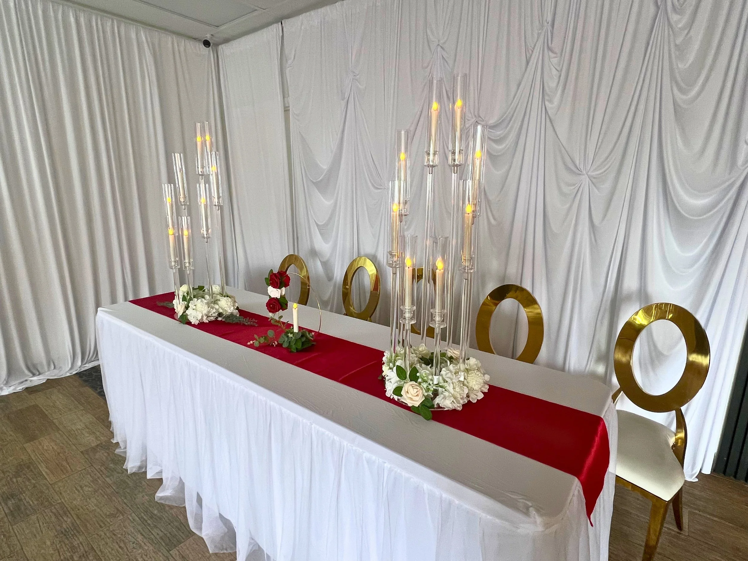 Elegant wedding reception table with white tablecloth, red runner, floral arrangements, and gold accents, set against white draped backdrop.