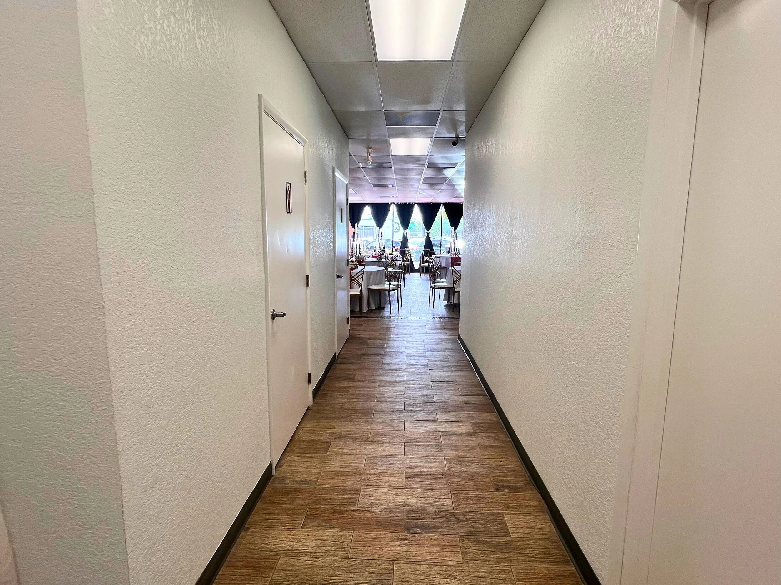 A hallway leading to a restaurant dining area with tables and chairs near windows with dark curtains.