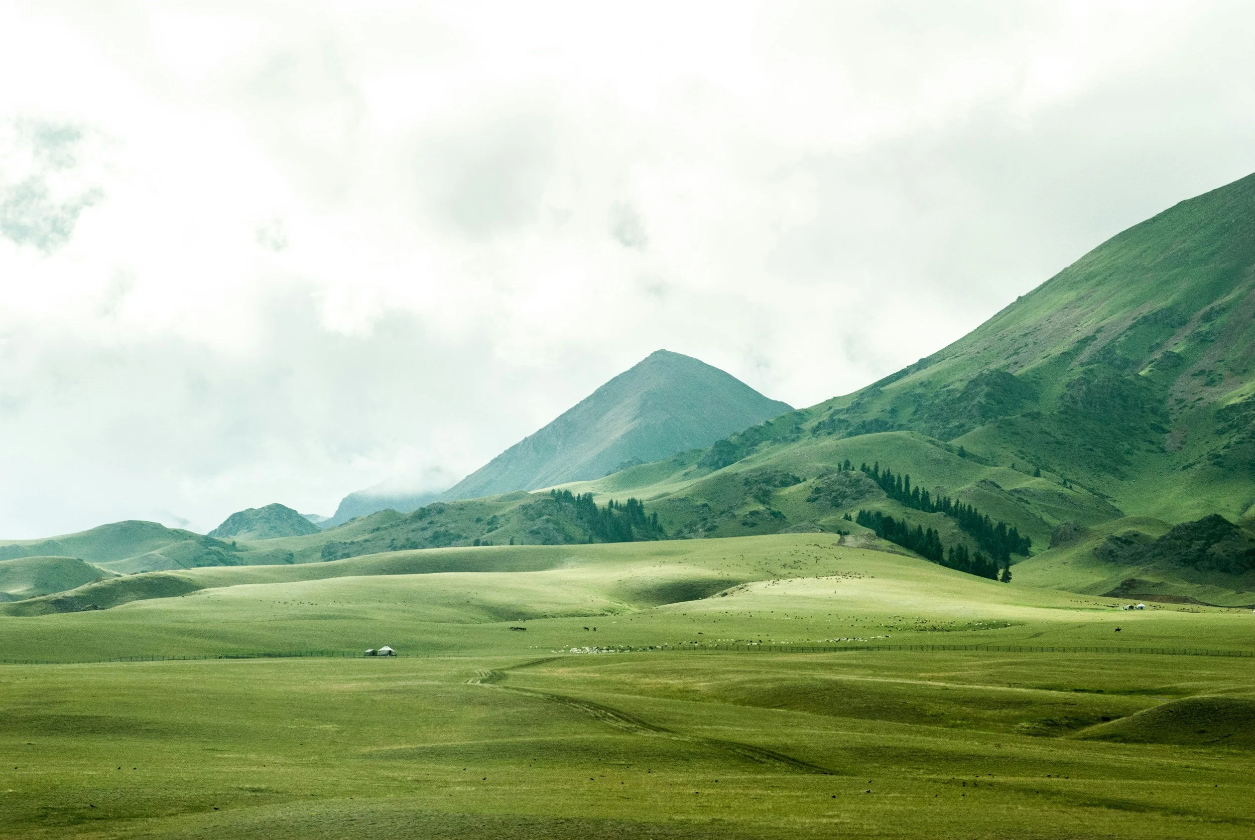 Green rolling hills and mountains under a cloudy sky with a few small structures visible in the distance.