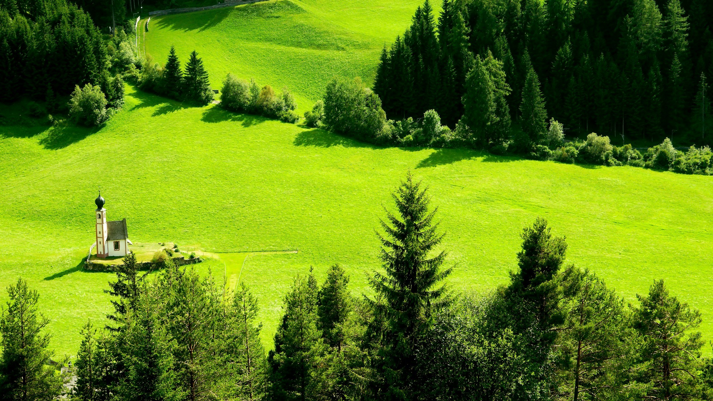 A small white church with a black onion dome sits on a grassy hill surrounded by green trees and rolling green fields in a rural landscape.