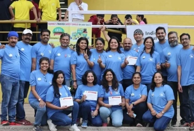 Group of people wearing blue shirts, some holding certificates, gathered outdoors with a yellow railing and a banner in the background.