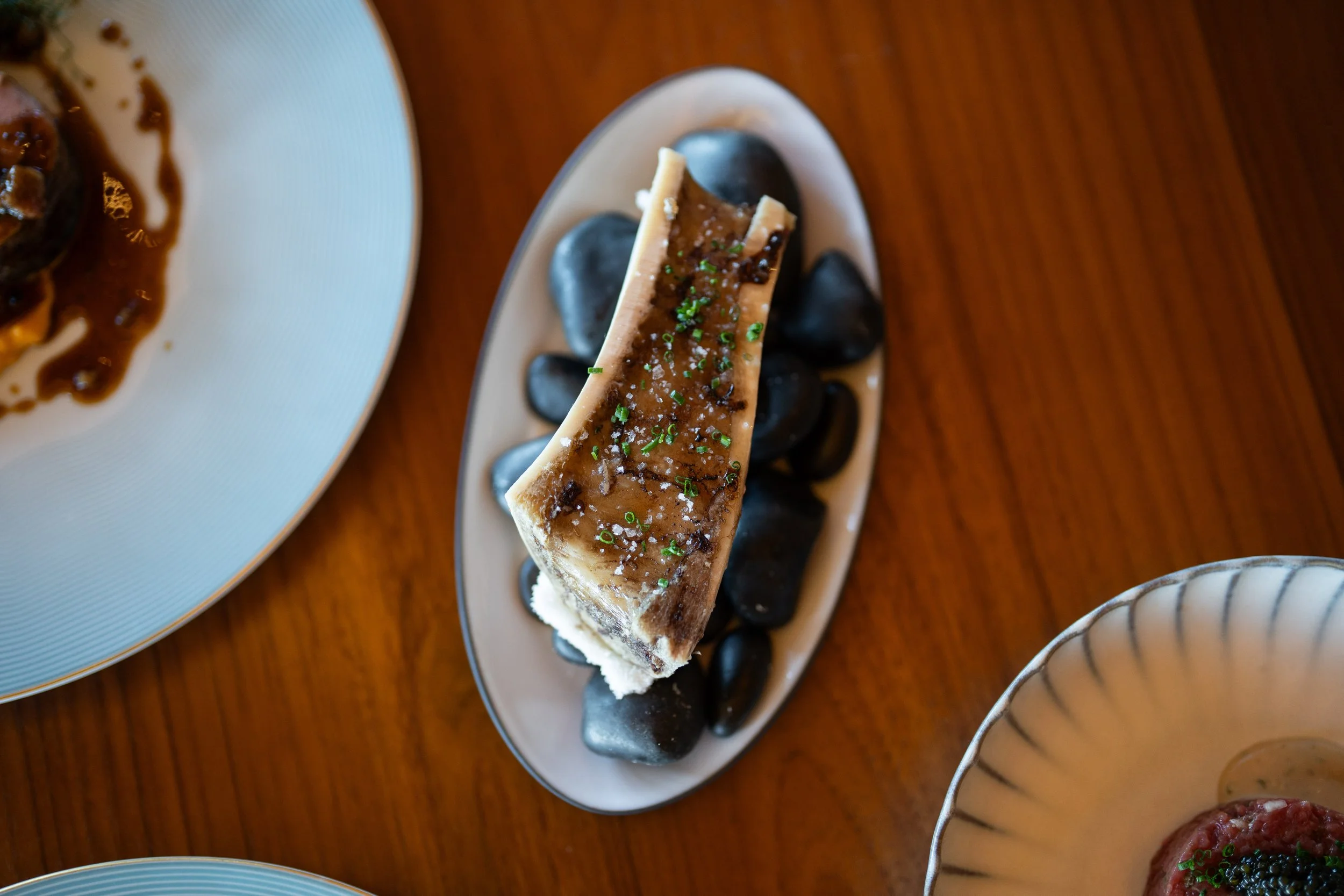 A slice of beef or marrow bone placed on a bed of black stones on an oval white plate, garnished with chopped green herbs and coarse salt.