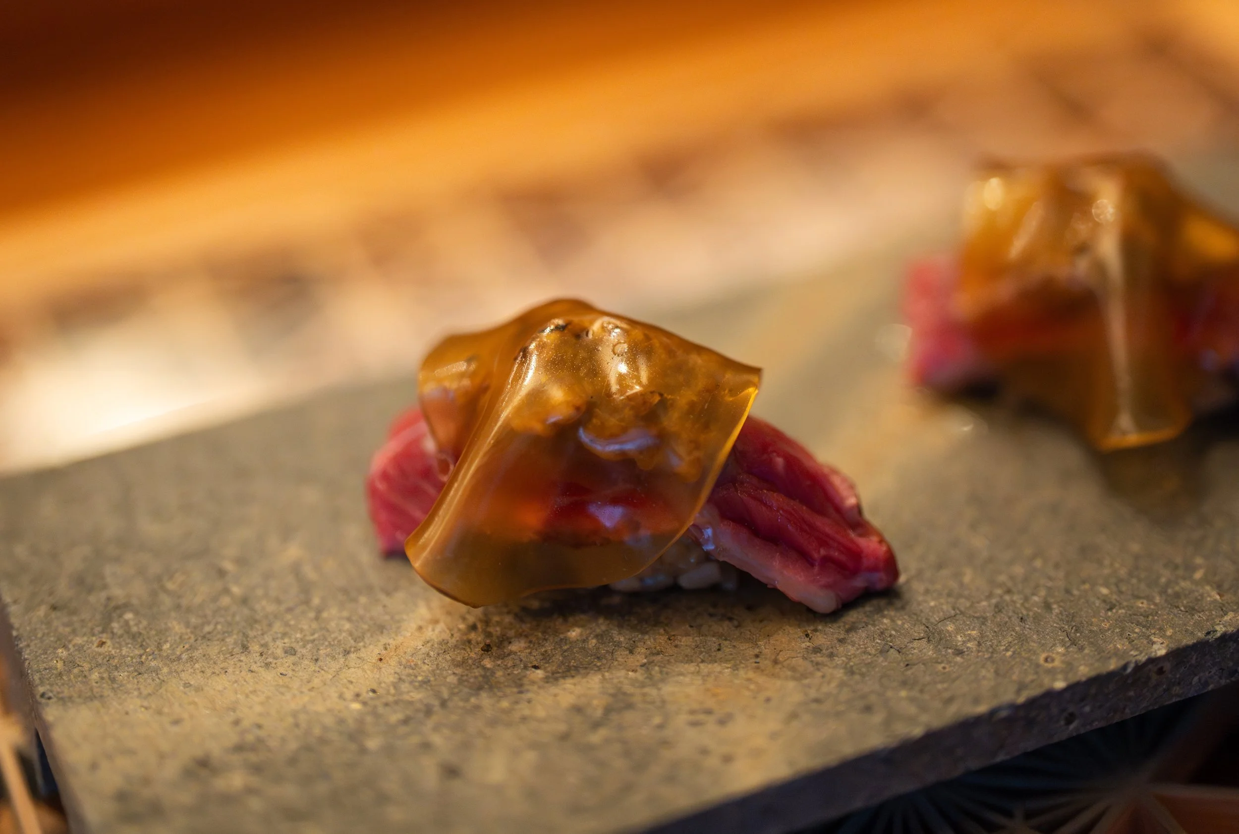 Close-up of a piece of sushi with raw beef, topped with a slice of caramelized ginger or mushroom on a stone serving plate.