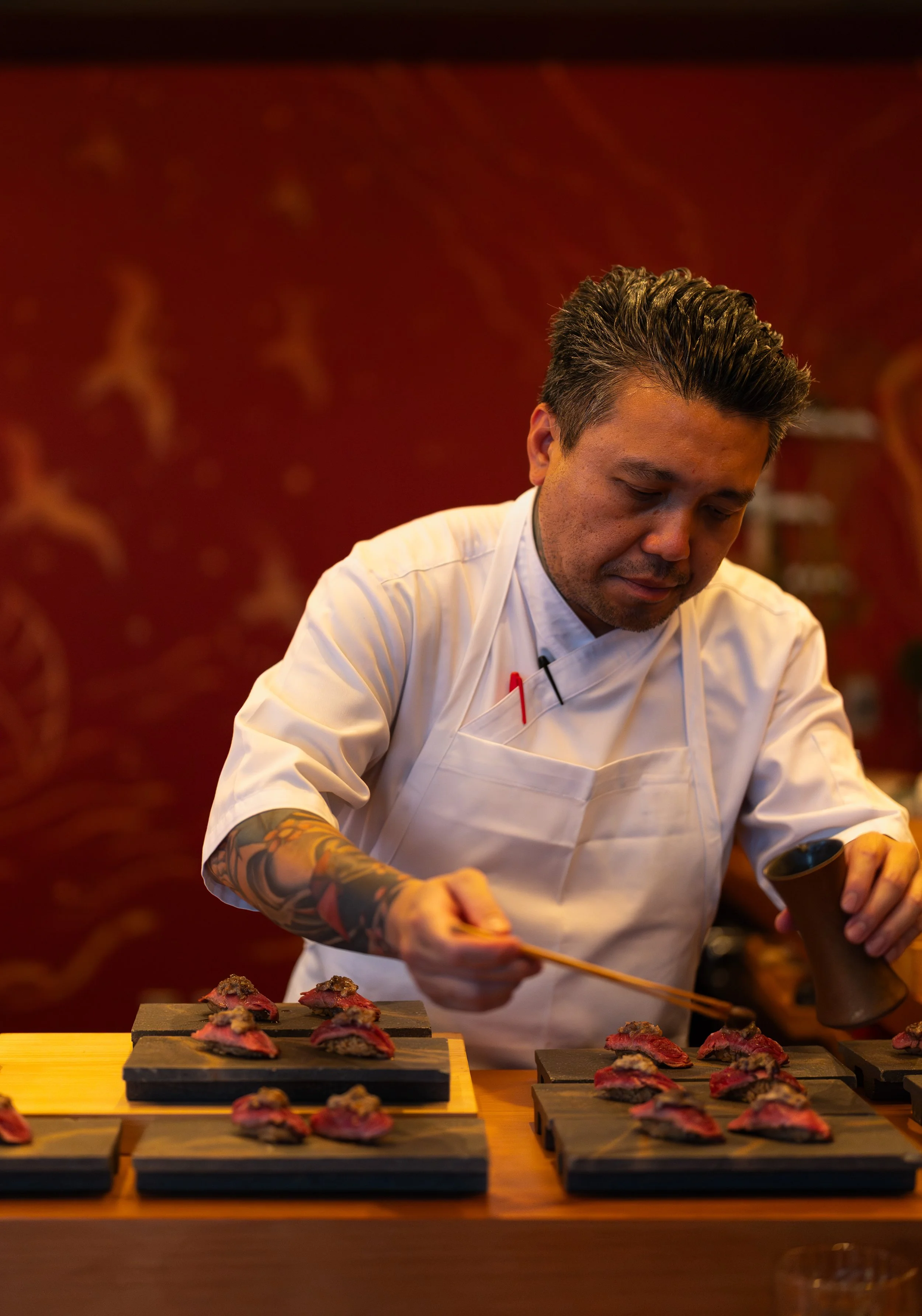 A chef with tattoos on his right arm arranging pieces of raw meat on black serving boards in a restaurant kitchen with red patterned walls.
