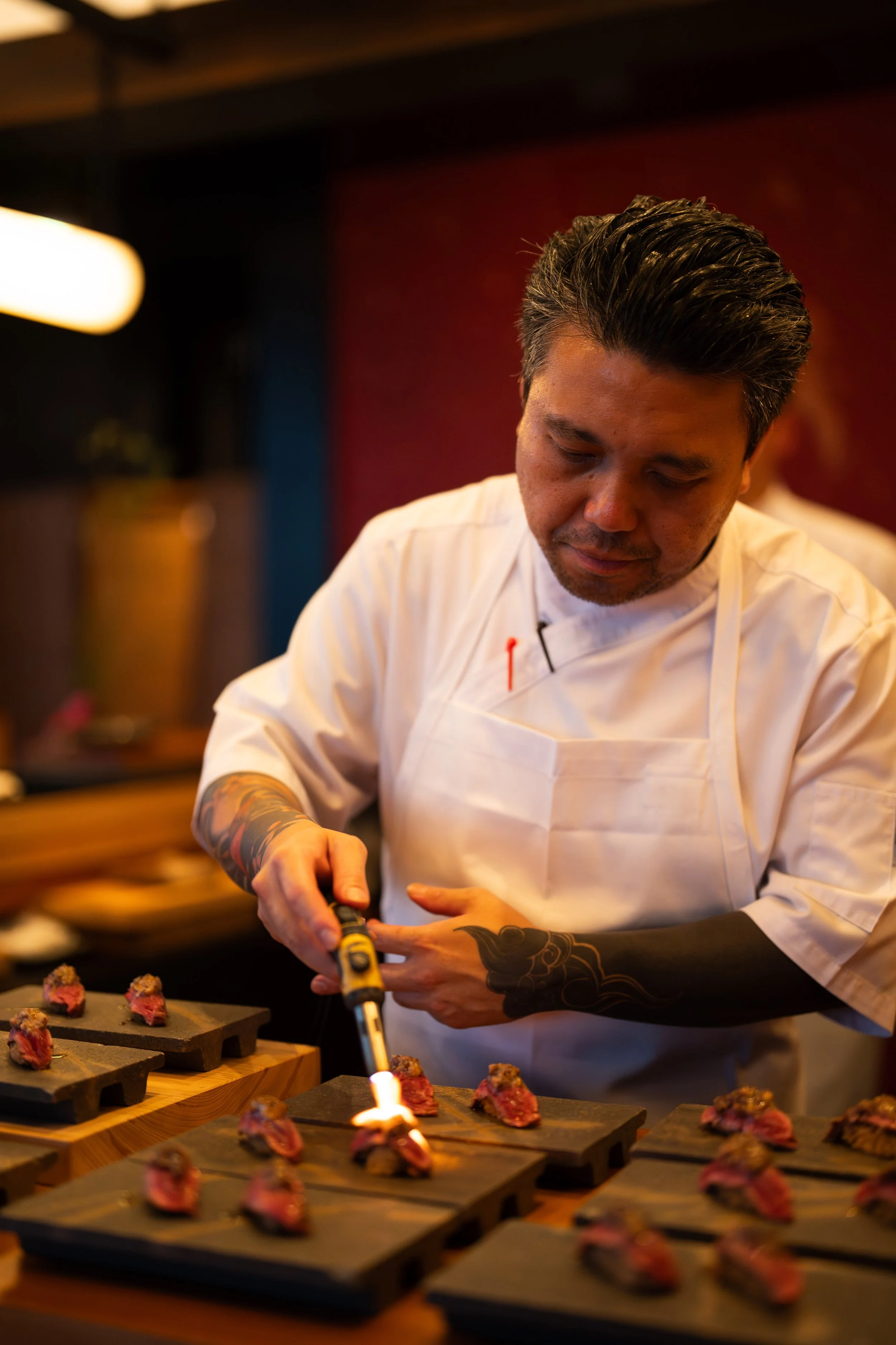 Chef using a torch to sear or add finishing touches to small portions of cooked meat on black rectangular plates in a restaurant kitchen.