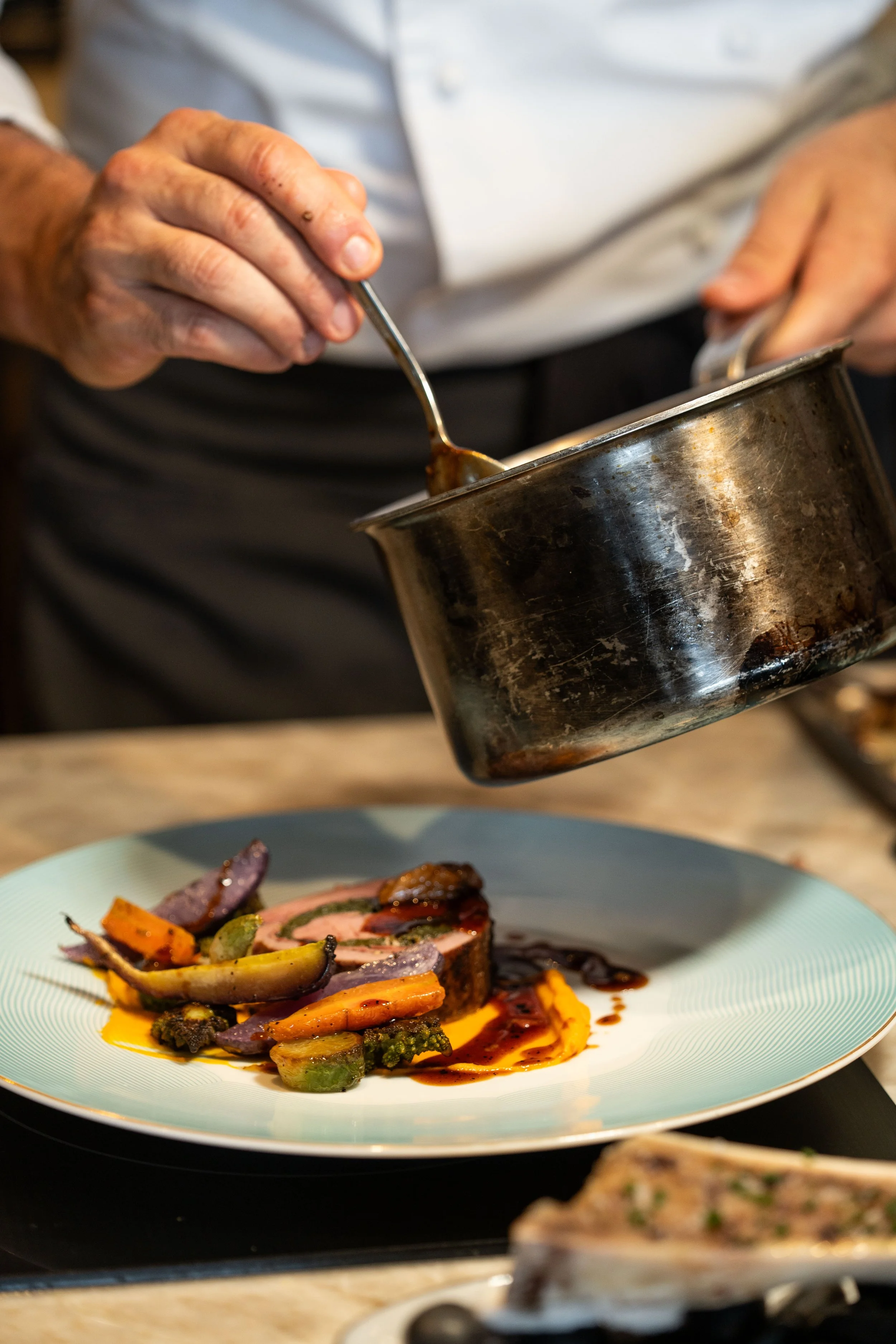 A chef is pouring a dark sauce onto a plated dish with roasted vegetables and a piece of meat. The focus is on the hand holding a spoon and the sauce being drizzled over the food.
