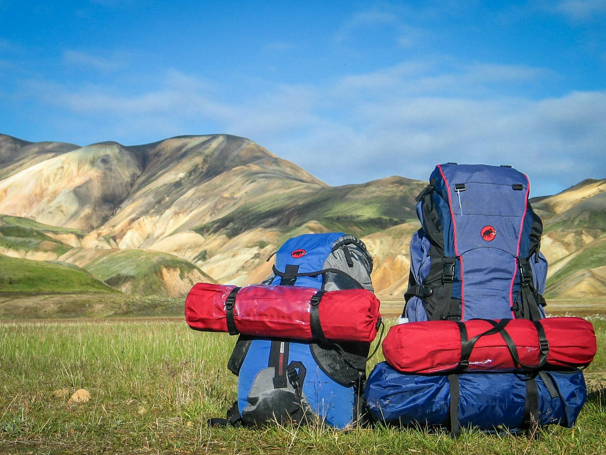 A collection of outdoor camping and hiking gear, including backpacks, tents, and sleeping bags, set against a mountainous landscape with rolling hills and a blue sky.