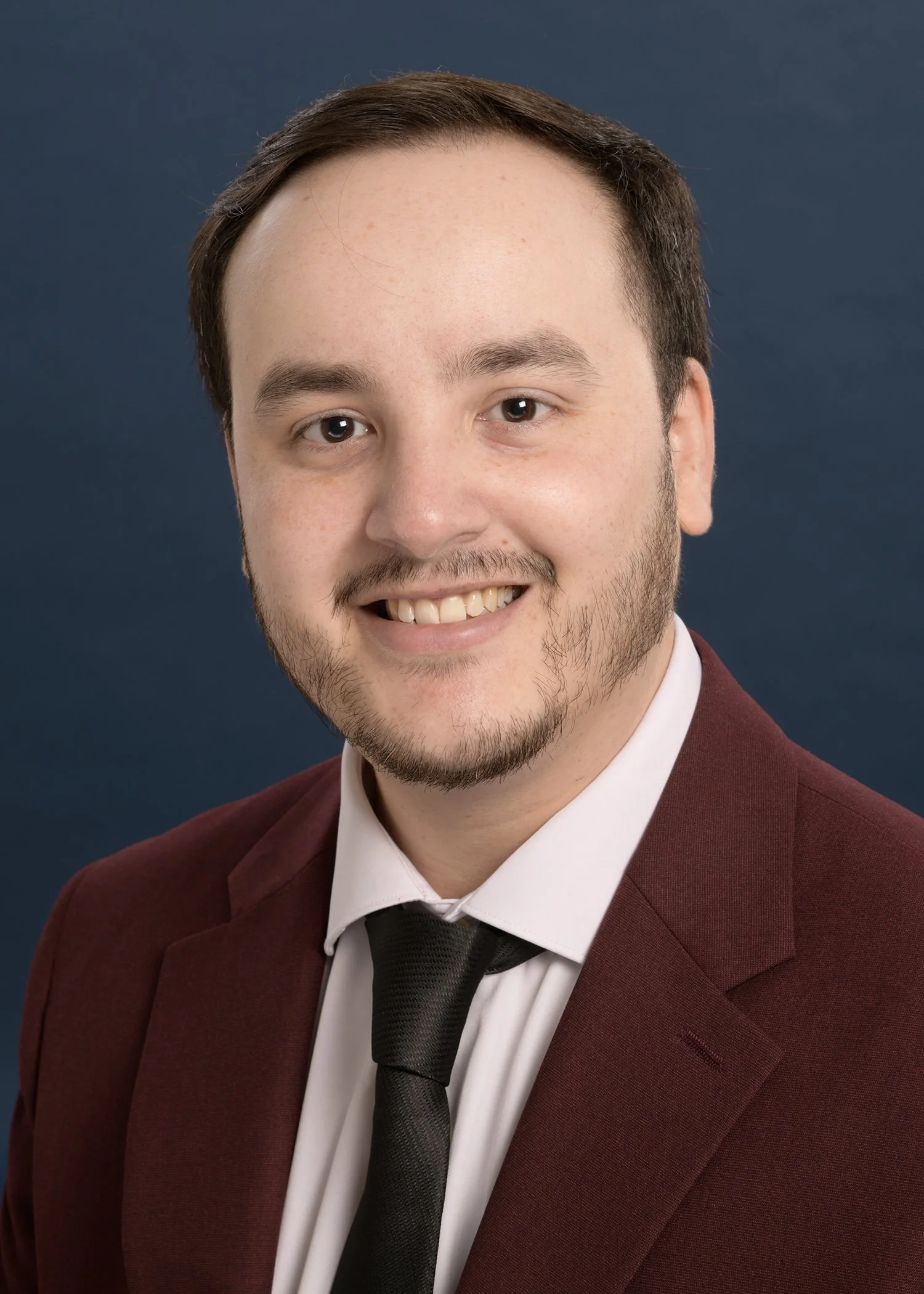 Young man with short brown hair, a beard, and a mustache, smiling, wearing a burgundy suit jacket, white shirt, and black tie, against a dark blue background.