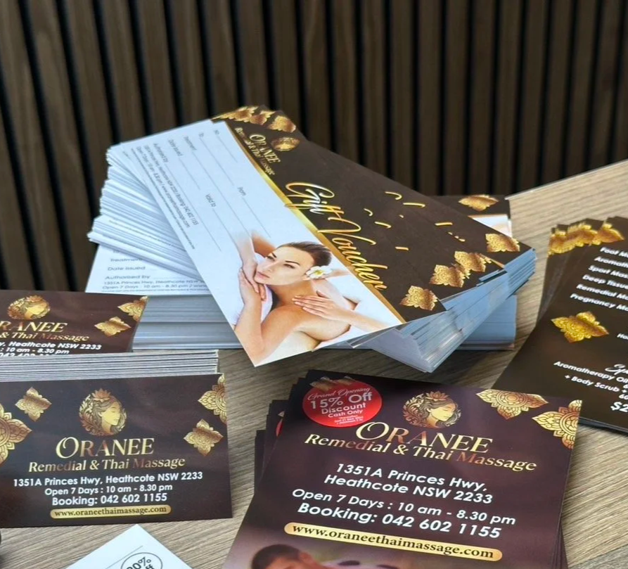 Stack of promotional flyers and business cards for ORANEEm Thai massage, showing contact information and promotional discounts, on a wooden surface.